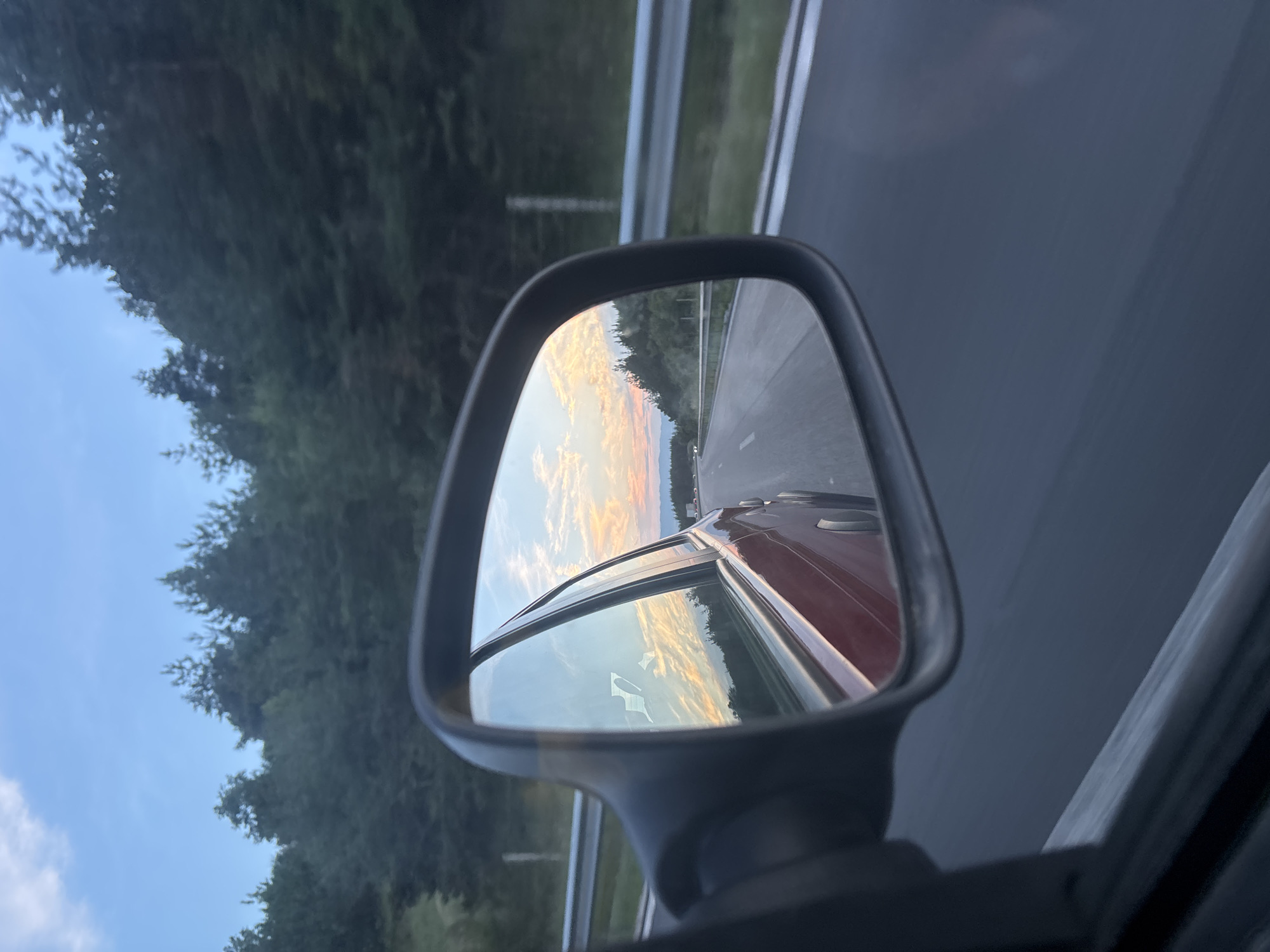 The image shows a rearview mirror reflection of a highway at dusk. The reflection displays a road with lane markings, a guardrail, and trees lining the side of the road. The sky in the reflection is filled with colorful clouds, suggesting a sunset. The original image captures a side view of a car, with the rearview mirror prominently in the foreground, showing the reflected scenery. The location is identified as Baliovtsi, Bulgaria.