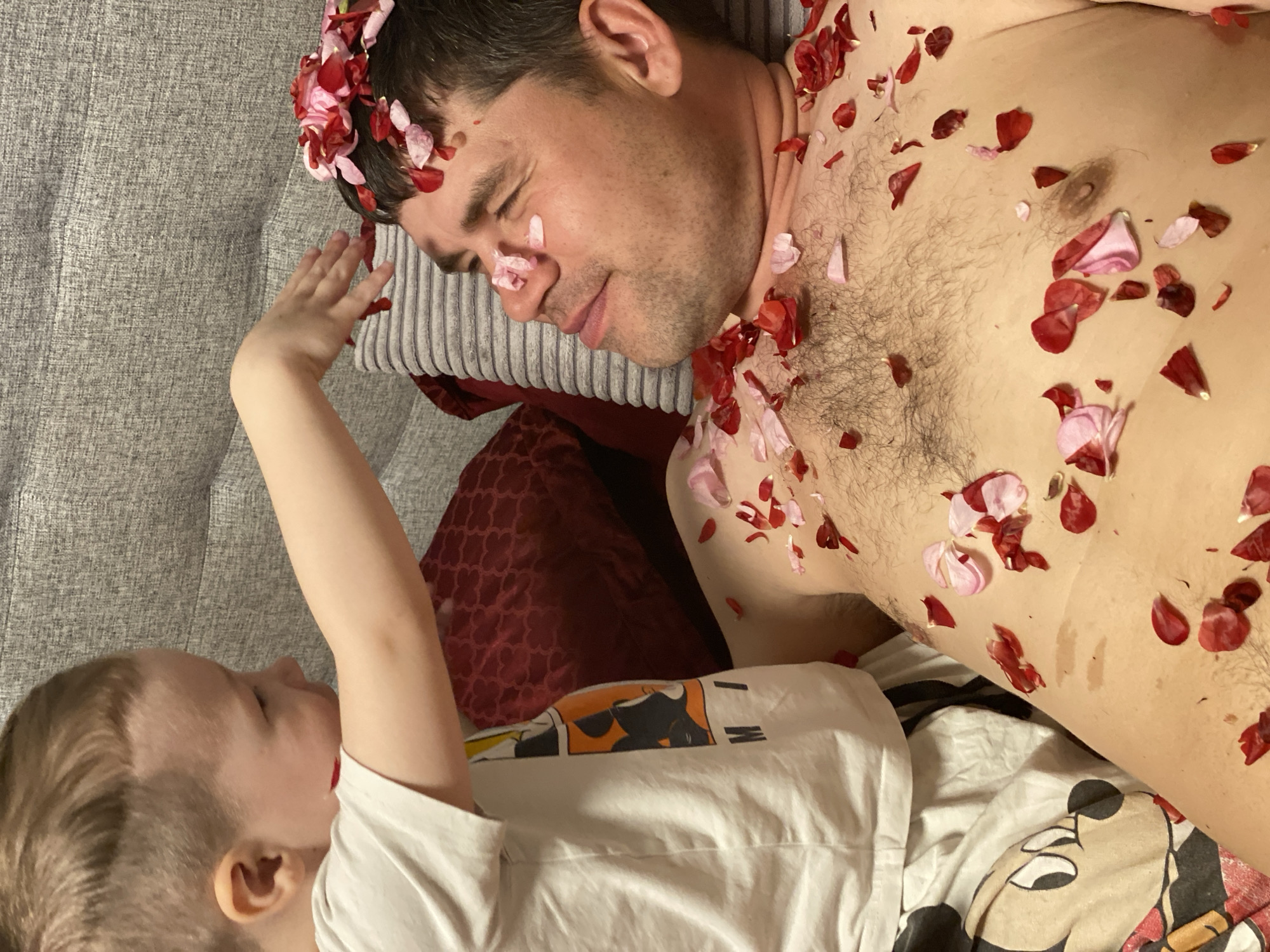 The image, taken in Sochi, Russia, depicts a tender and playful indoor scene between an adult male and a young child. The adult male lies on his back on a grey fabric couch or bed, eyes closed, suggesting rest or sleep. He is bare-chested with visible body hair and stubble, and is covered with numerous red and pink rose petals scattered across his chest, shoulders, head, and face, including some on his nose. A young child, with light hair and wearing a white t-shirt featuring a cartoon character (partially visible with the text "I'M"), is positioned to the left of the adult. The child extends a hand towards the adult's head, seemingly engaged in placing or touching the petals. A deep red patterned pillow supports the adult's head, and a Mickey Mouse themed bedsheet or blanket is visible beneath the child. The scene conveys a relaxed, affectionate, and perhaps humorous interaction between the two individuals.