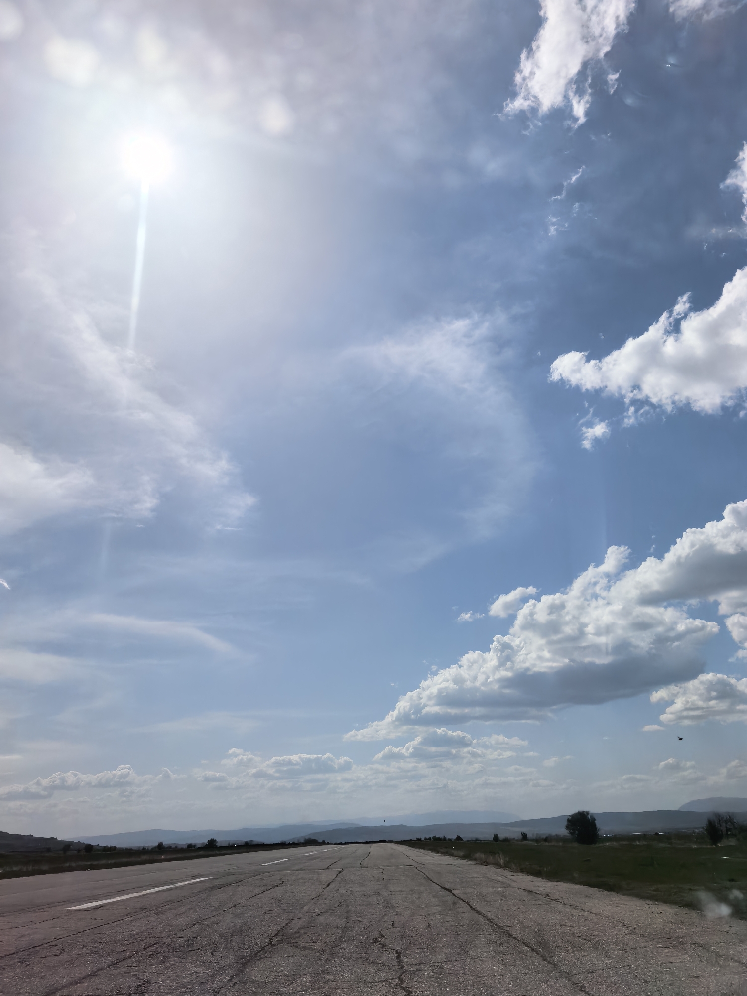 The image displays a wide-angle perspective of a rural landscape, dominated by a vast sky. A long, cracked asphalt road, marked with a faded white line on its left, stretches from the foreground into the distance, appearing to crest a slight rise. On either side of the road, the land consists of dry, light green fields with scattered bushes and trees. In the far distance, a series of muted blue hills or mountains softly delineate the horizon.
The sky, which occupies the majority of the frame, features a brilliant, intensely bright sun in the upper-left quadrant, producing a strong lens flare and a distinct sunbeam. Scattered white cumulus and stratocumulus clouds populate the expansive blue sky, varying in size and density, with some appearing particularly radiant from the direct sunlight. A small, dark bird is visible in flight against the blue sky in the upper-right portion of the image.
The scene suggests a clear, bright day, likely midday or early afternoon, given the high position and intensity of the sun. The overall impression is one of open space, journey, and a serene, unpopulated environment. There are no visible people, specific activities, or discernible text.