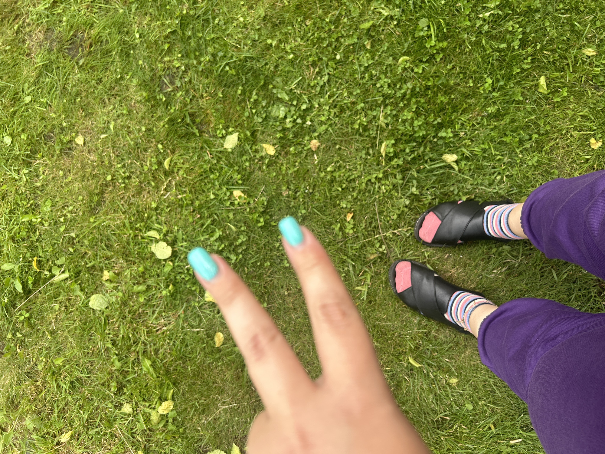 The image shows a pair of legs and feet from a person's perspective, sitting on grass. The person is wearing purple pants and black slip-on sandals with pink accents. They are also wearing socks with horizontal stripes in pink, white, and blue. One hand is visible, making a peace sign with brightly painted turquoise fingernails. The background is a grassy lawn with some fallen leaves, suggesting an outdoor setting. The lighting indicates it is daytime, likely a clear day given the sharp shadows. There is no visible text in the image. The overall mood appears casual and relaxed, with the person possibly enjoying a moment outdoors in Leningradskaya, Russia.
