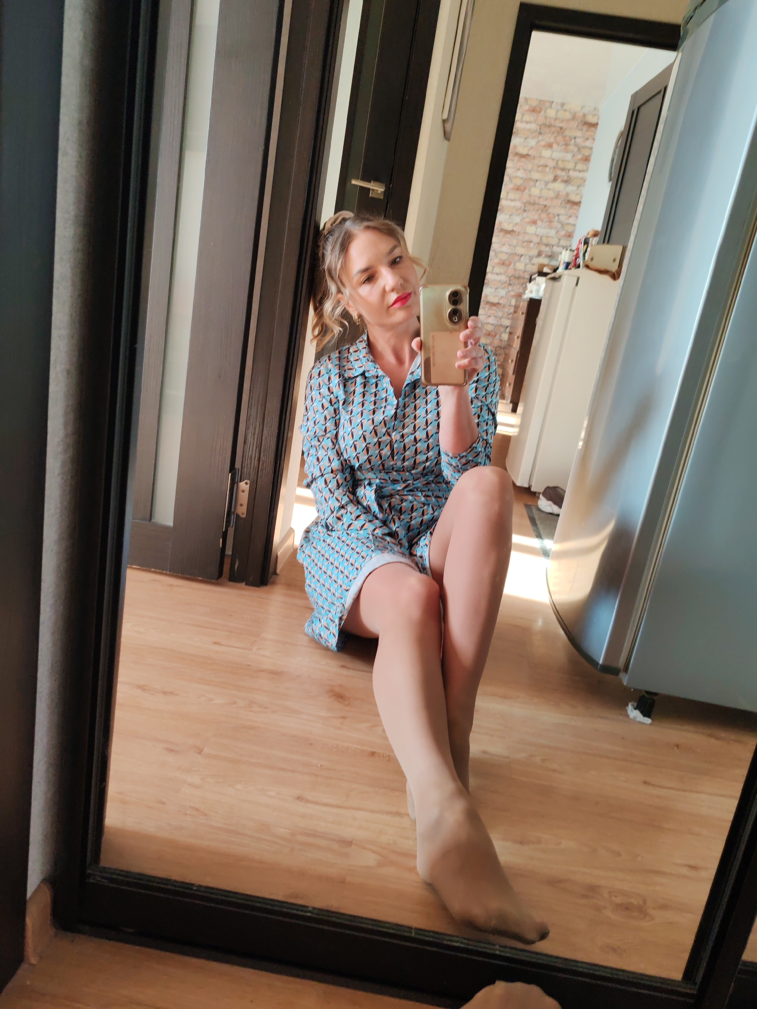 A woman is sitting on a wooden floor in what appears to be an apartment, taking a selfie in a mirror. She has fair skin, blonde hair tied back, and is wearing a patterned blue and brown dress with nude-colored stockings. She is holding a smartphone in her right hand, with the back of the phone facing forward.
Behind her is a doorway with a dark-framed door. Through the doorway, a room is visible with a brick wall and a glimpse of white furniture. To the right of the woman is a stainless steel refrigerator.
The scene is indoors, well-lit, and appears to be a casual, everyday moment. There are no immediate indications of the location being in Petropavlovsk-Kamchatsky, Russia; the setting could be in any similar urban apartment.