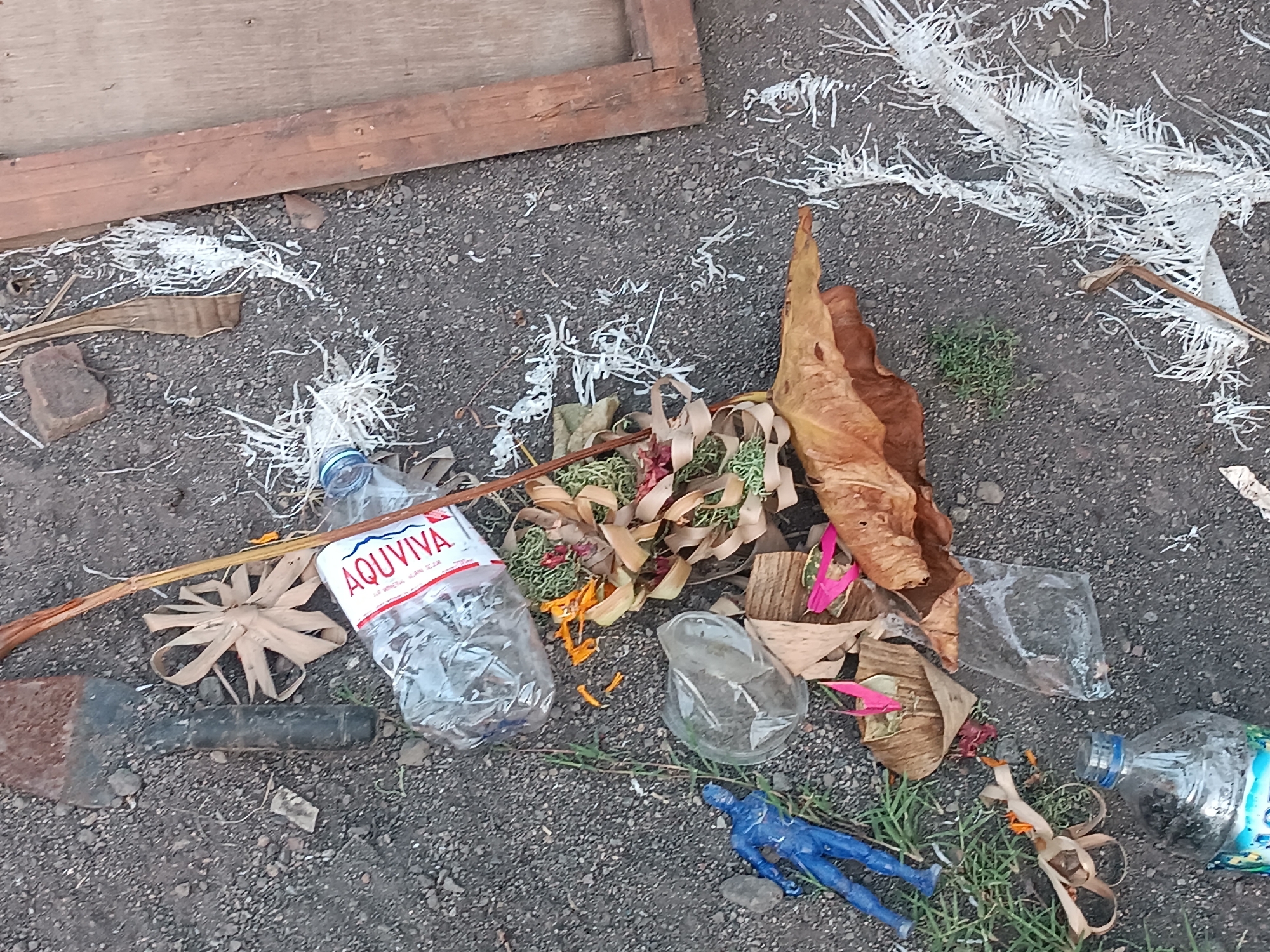 This image depicts a collection of discarded items and debris on a gravelly surface, possibly indicating a roadside or outdoor area. In the upper left quadrant, a portion of a wooden structure, likely a fence or panel, is visible. Scattered across the ground are various objects, including a plastic water bottle with the label "AQUVIVA" clearly visible, a broken glass bottle or container, dried leaves and foliage, and what appears to be a blue plastic figurine resembling a person. There are also remnants of what might be some form of offering or ritualistic items, such as small colorful petals and woven plant material. A metal tool, possibly a spade or hoe, lies horizontally in the lower left portion of the image. White, fibrous material is strewn across the ground, mixed with the gravel and other debris. The overall scene suggests a neglected or recently disturbed area. The scene is likely located in Tabanan, Indonesia, given the context provided.