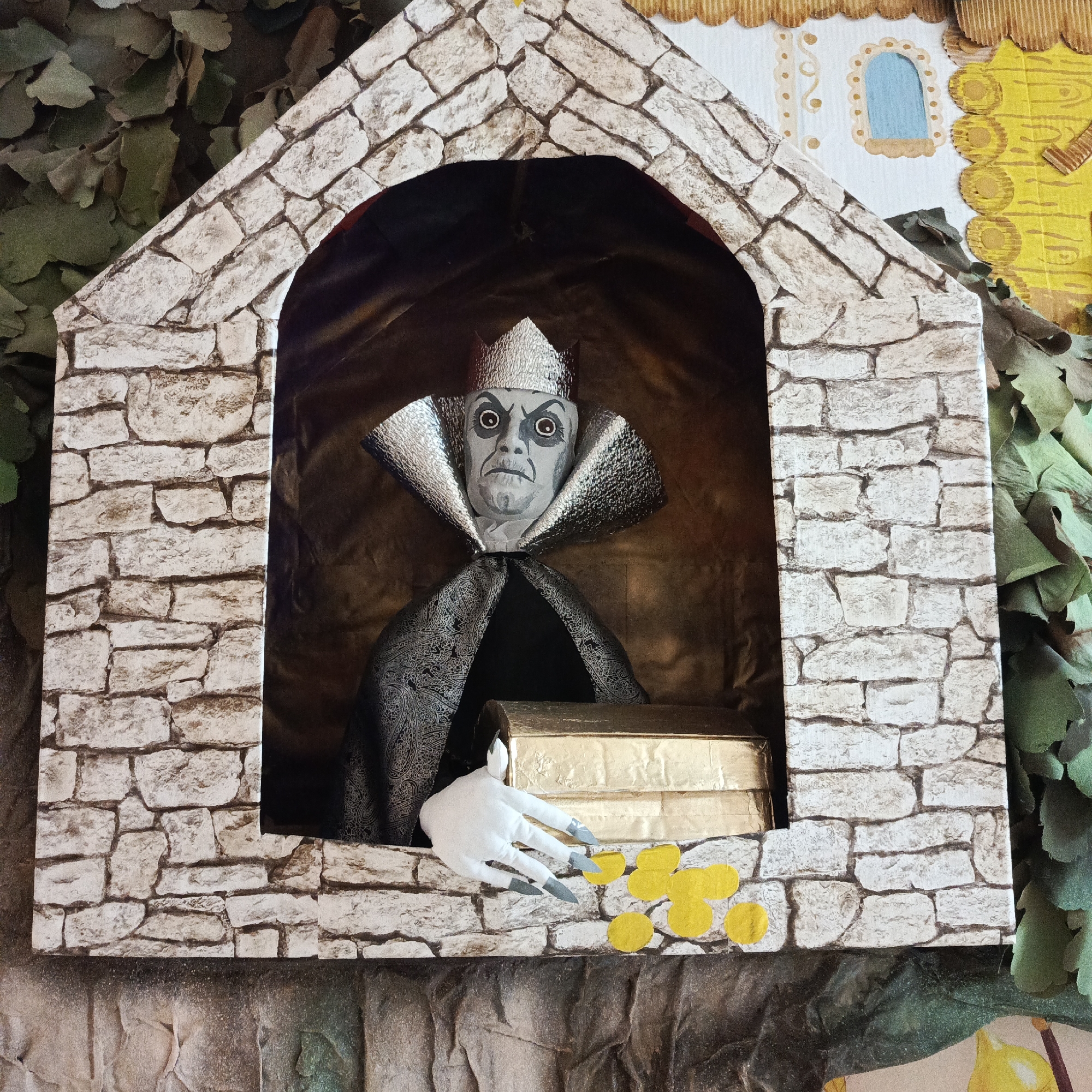 The image shows a handmade diorama of a scene featuring a sinister-looking character in a stone structure resembling a castle facade. The figure is gray-skinned with a silver crown and a wide, patterned collar, wearing a black cape. It has a gold chest in front of it, and a white, clawed hand is reaching towards yellow, circular objects, possibly representing coins. The stone structure is made of cardboard with a printed stone pattern. The background behind the figure is brown and possibly fabric. The setting seems to be an outdoor area, with greenery, possibly artificial leaves, around the structure. Based on the details, it's difficult to confirm the specific location as Markovskii, Russia without further context or information associated with the image. The scene is reminiscent of a fairy tale or fantasy setting with a villainous character.