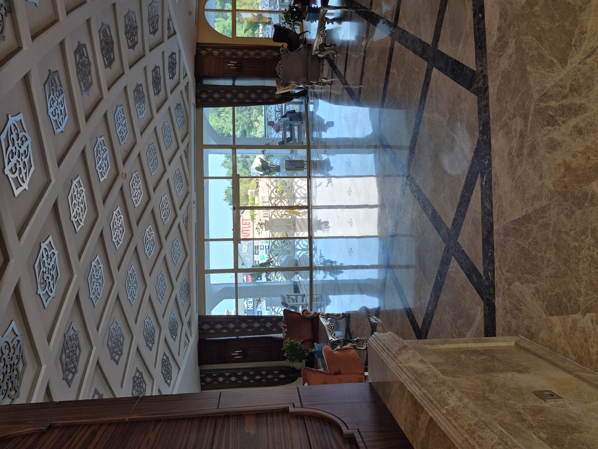 This indoor shot shows the interior of a building, possibly a hotel or a high-end shop. The scene is in Alanya, Turkey, as indicated by the "OUTLET" sign visible through the large windows. The interior is opulent, with a marble floor featuring a geometric pattern. The ceiling is decorated with square panels, each containing an ornamental design. Large windows provide a view of the outdoors. The furniture includes ornate chairs and tables, giving the area a formal feel. There are no visible people in the foreground. The lighting is bright, likely due to the natural light coming through the windows.