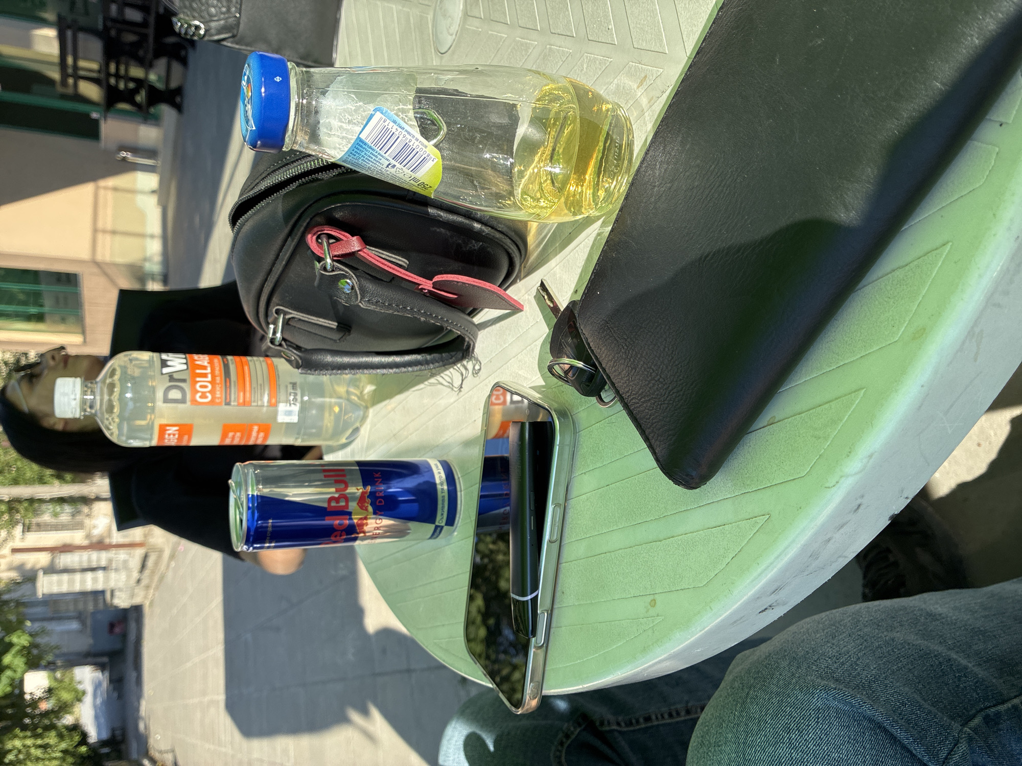 The image depicts an outdoor setting, likely a cafe or public seating area, with a light green plastic table in the foreground. On the table, a silver can of Red Bull, a bottle of "Dr.Wi COLLAGEN" energy drink, a glass bottle containing a light yellow liquid, a black leather pouch with pink accents, a smartphone with a vape pen resting on it, a black leather wallet, and a set of keys are arranged. A person's legs, clad in blue denim jeans, are visible on the left side of the frame, suggesting they are seated at the table. In the background, a person wearing a black top is partially visible, looking away from the camera. The environment suggests a sunny day with trees and buildings in the background, characteristic of an urban or semi-urban area. The presence of multiple beverage containers and personal items indicates a casual gathering or a break. Based on the context provided, this scene is located in the city of Aglen, Bulgaria.