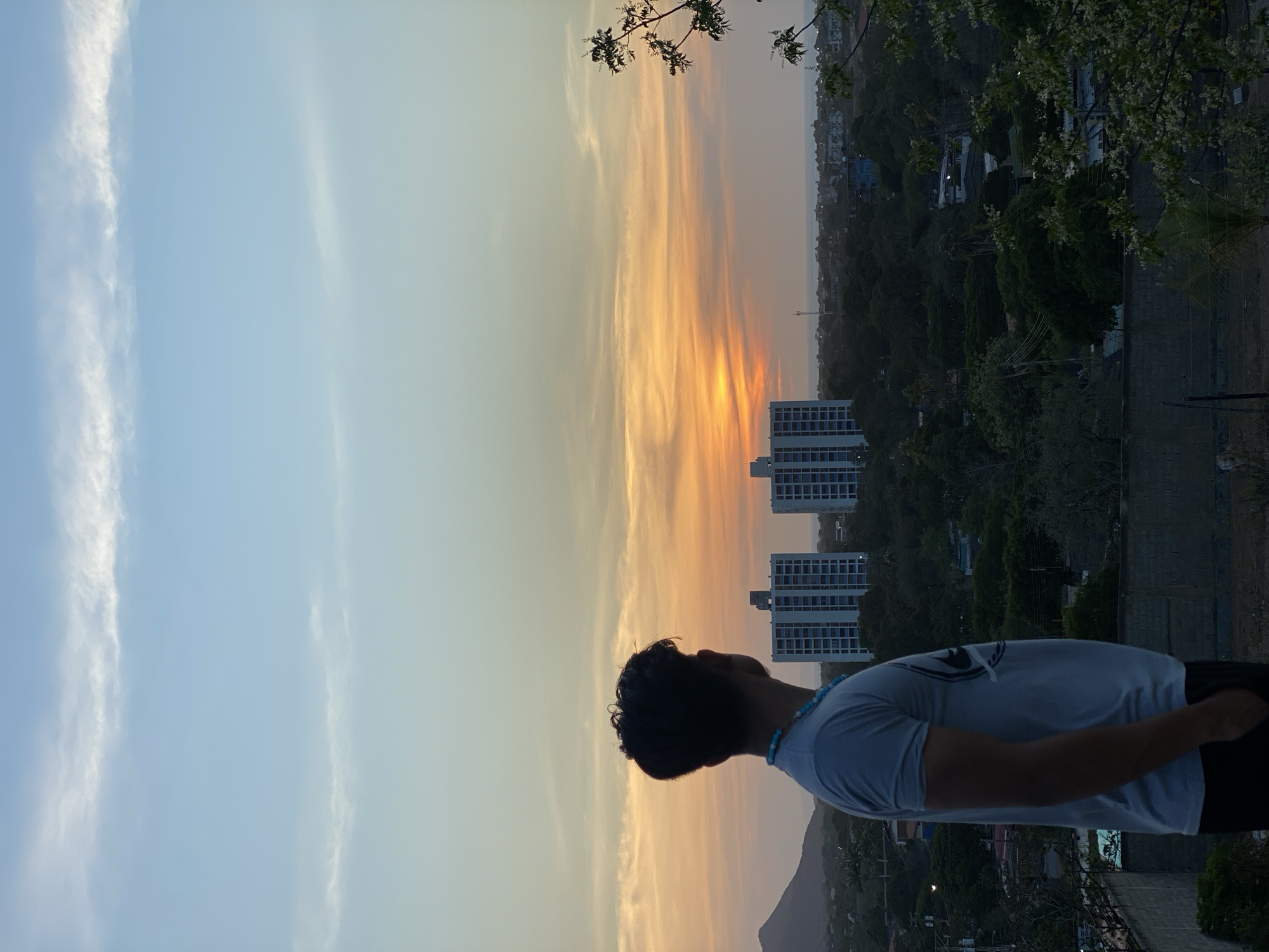 A man stands silhouetted against a sunset sky in Juan Griego, Venezuela. He faces towards the horizon, where the sun casts an orange and yellow glow amidst wispy clouds. The sky transitions to a pale blue on the left side of the image. In the background, a few tall buildings are visible, along with a lush green landscape dotted with trees. The scene appears to be shot from an elevated position. The man is wearing a white T-shirt and a blue beaded necklace.