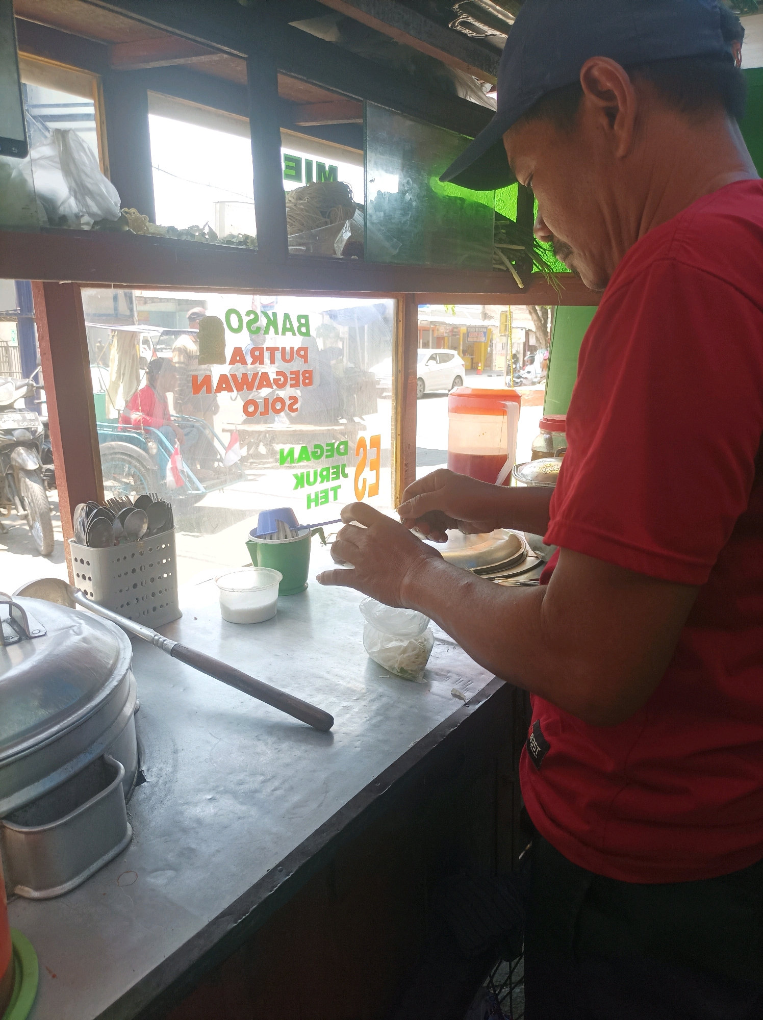 A man wearing a red shirt and a dark blue cap is standing behind a metal counter in what appears to be a food stall or small restaurant. He is in the process of preparing food, with his hands actively engaged in some task. On the counter, there are various items, including a metal container with cutlery, a small plastic container, and a larger metal pot with a lid. A red liquid is visible in a large plastic dispenser behind him. Through a window behind the counter, we see an outdoor scene with a street, vehicles, and buildings, suggesting an urban or semi-urban environment. Text on the window, possibly the name of the establishment, is partially visible and reads "MIE" and "BAKSO PUTRA BECGAVAN SOLO ES DEGAN TEH". The lighting suggests it is daytime, with sunlight streaming in, creating some glare on the window. The setting is consistent with a street food vendor in Sumuragung, Indonesia.