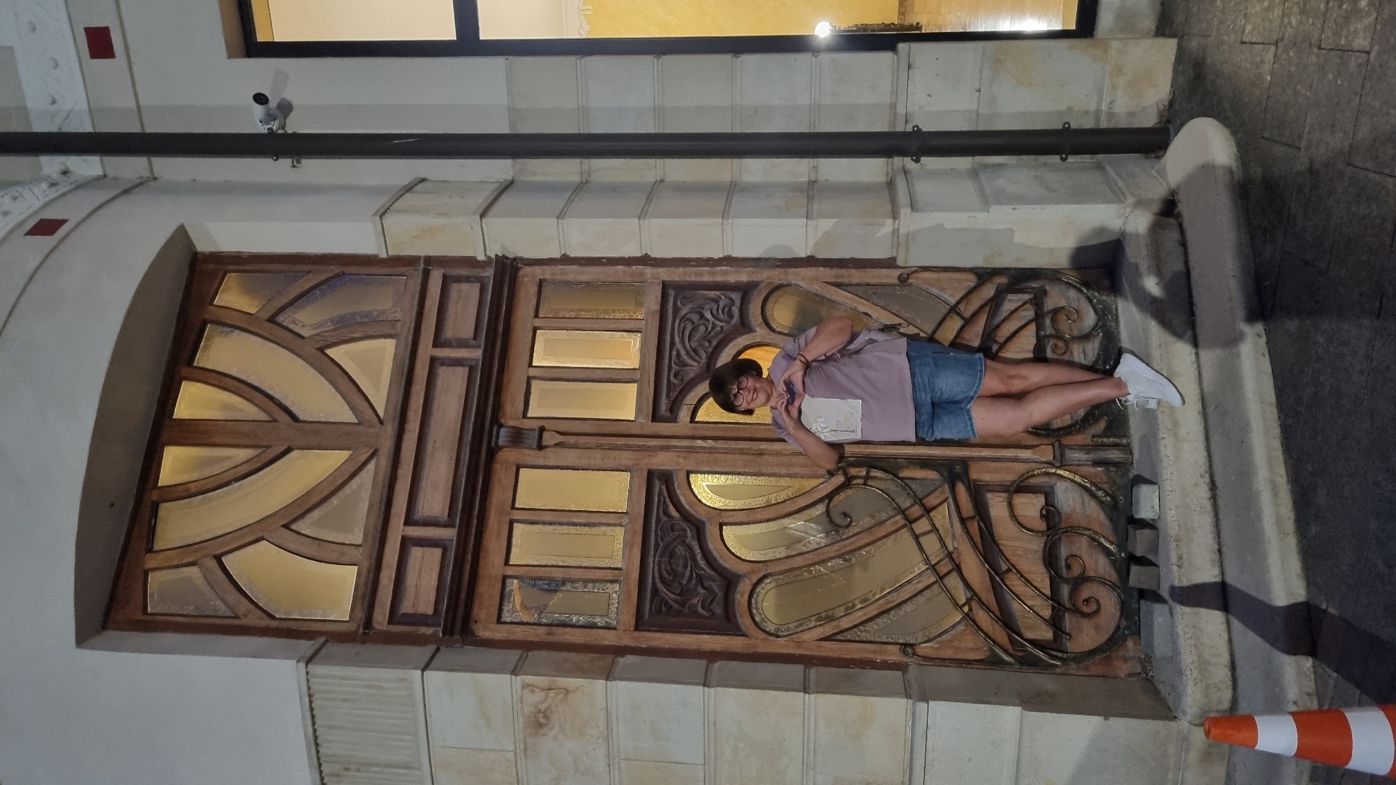 A woman stands in front of a large, ornate wooden door with glass panels in the city of Berehove, Ukraine. She is wearing a pink t-shirt, denim shorts, and white sneakers. She is making a heart shape with her hands over her chest and smiling at the camera. The door is flanked by stone columns and is illuminated by interior lights, casting a warm glow. The scene appears to be at night or in low light conditions, with shadows cast on the ground. A traffic cone is visible in the lower left corner of the image. The architecture suggests a historic building, possibly a cultural institution or a former grand residence.