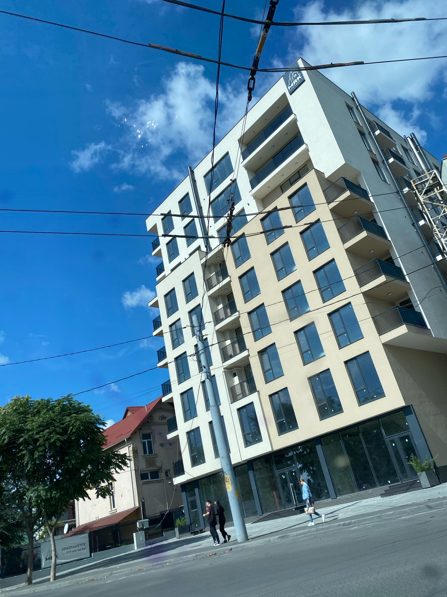 A modern, cream-colored apartment building with multiple balconies and dark-paned windows is situated on a street in Chișinău, Moldova. The building has a logo with "MRK" and what appears to be a house icon at the top. To the left of the modern building, an older, lighter-colored building with a red roof is visible. A lush green tree stands in front of the older building. The street in front of the buildings is paved, and a sidewalk runs alongside it. Two people, one in black and another in dark clothing, are walking on the sidewalk in front of the modern building. Further down the sidewalk, another person in a blue patterned shirt and jeans is walking. Overhead, power lines are strung across the sky, which is a vibrant blue with scattered white clouds. There are some bright, reflective spots in the sky, possibly from light reflecting off the clouds or from a distant source.