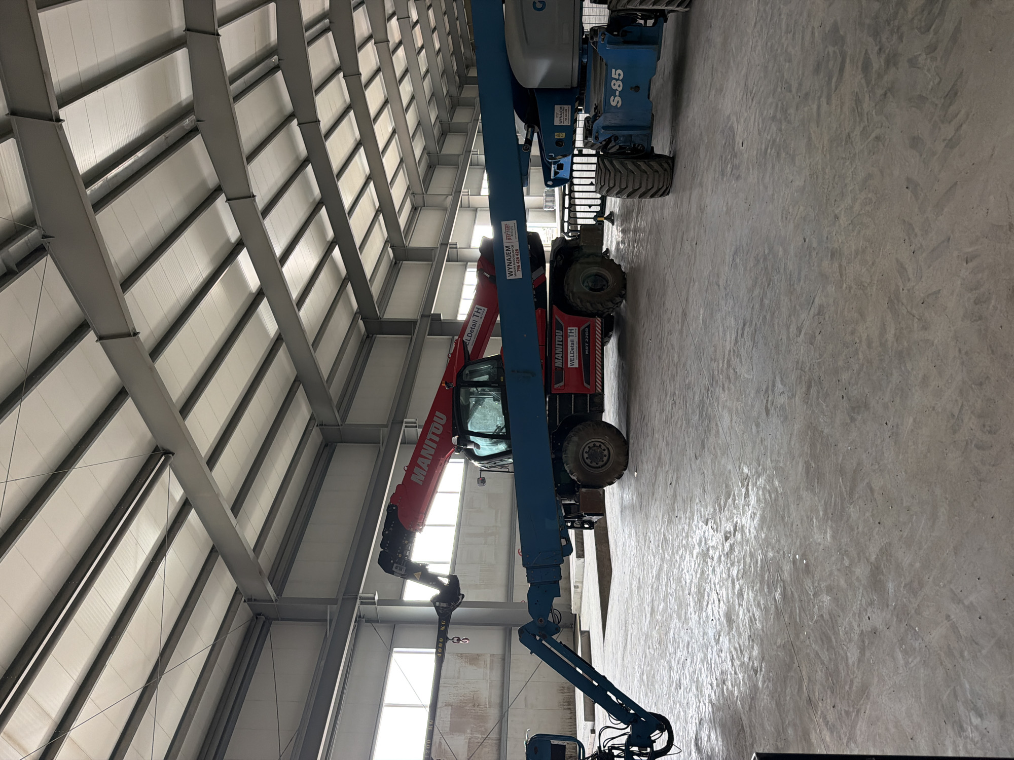 The image depicts the interior of a large, unfinished industrial building. The structure features a metal frame with a white insulated roof. In the foreground, several heavy-duty construction machines are parked on the concrete floor. On the left is a blue boom lift with its arm extended. To its right is a red Manitou telescopic handler with its boom extended and a hook attachment. Further to the right is a blue Genie boom lift, labeled "S-85." The floor is concrete and appears wet in places, reflecting the light coming from large windows on the far wall. The overall impression is a construction site within a newly built facility. The text "WYNTAJM 736 426 436" is visible on both the red and blue boom lifts, suggesting a rental company. The location of Janowice, Poland, is mentioned, but there are no visual cues in the image to confirm this specific location. No people are visible in the image.