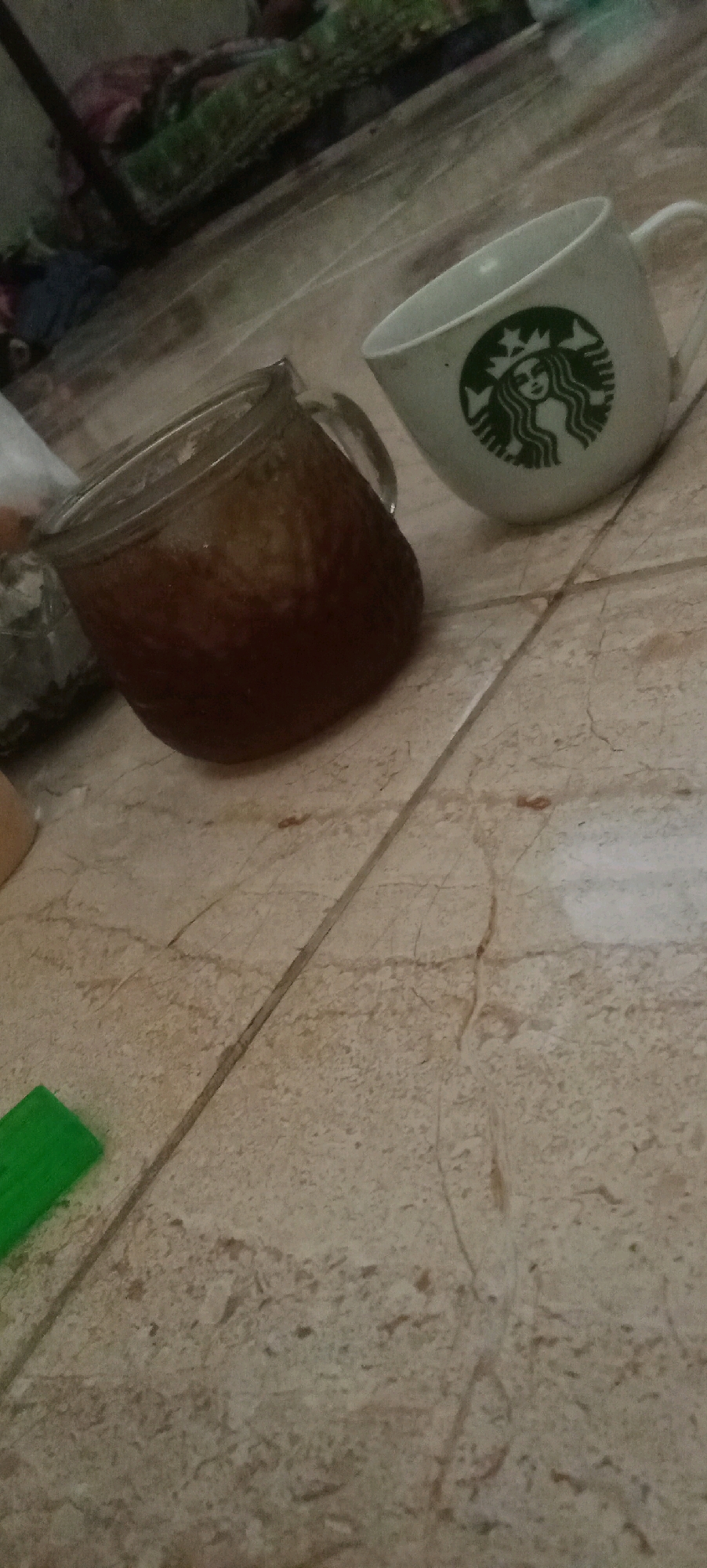 This is a close-up, low-angle, medium shot of two beverage containers on a tiled floor. In the foreground and slightly to the left is a glass mug filled with a dark liquid, possibly iced coffee or tea, with ice cubes visible. The mug is tilted, with the liquid filling about three-quarters of its volume. To the right and slightly behind the glass mug is a white ceramic mug with the green Starbucks logo on it. The logo depicts a siren. The background shows a blurry collection of objects on what appears to be a shelf or rack, including fabric items and possibly a metallic support structure. A small, bright green rectangular object, possibly a piece of plastic or a marker, is visible in the lower left corner of the frame. The floor is composed of beige tiles with visible grout lines and some texture. The lighting is dim, creating shadows and contributing to the overall moody atmosphere of the image. The scene is indoors, likely in a residential or casual setting. There is no identifiable information to suggest the specific location within Karawang, Indonesia.