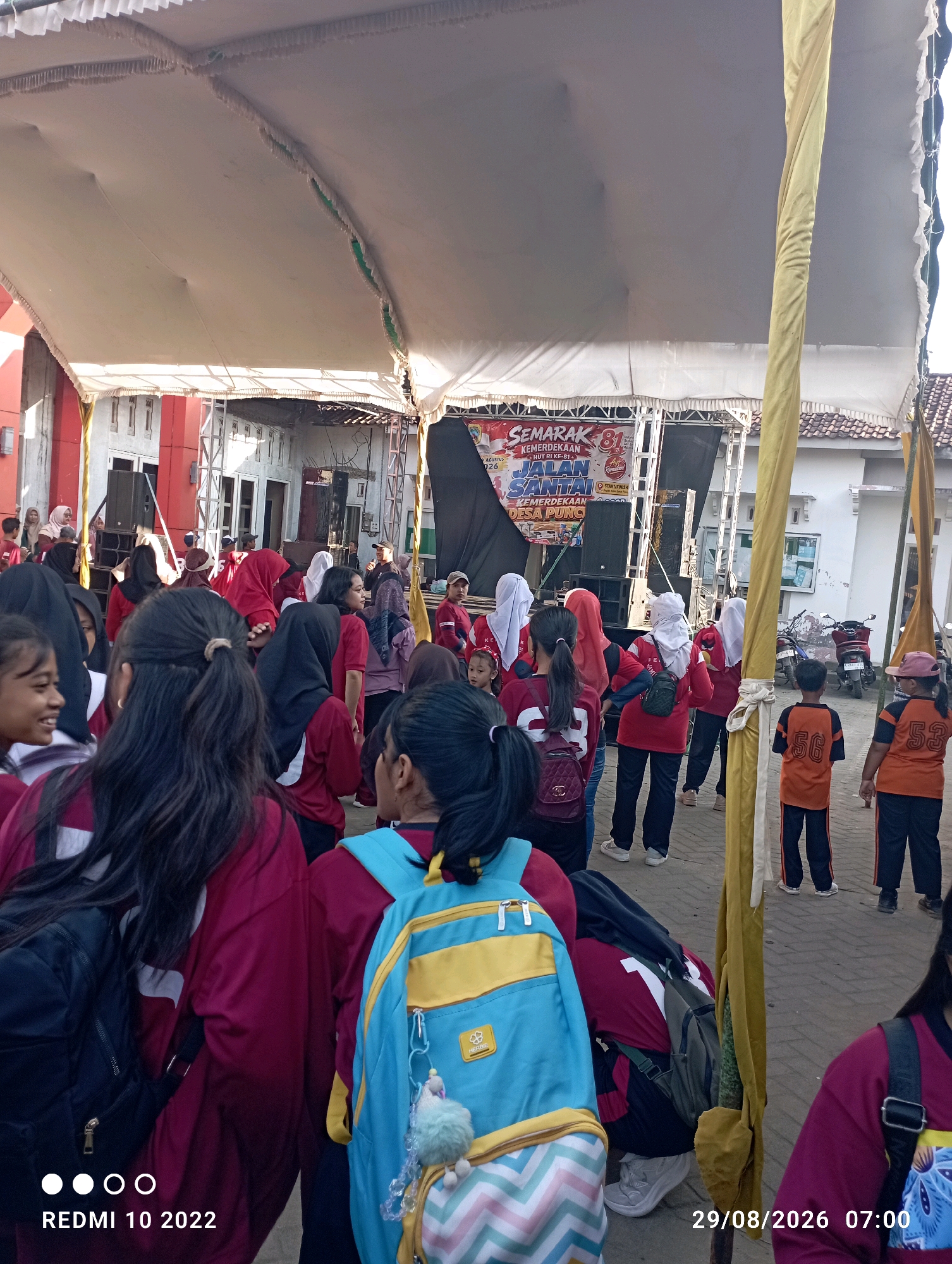 The image depicts an outdoor event taking place under a large tent, likely in Dukuhseti, Indonesia. A crowd of people, predominantly young individuals, are gathered. Many are wearing matching maroon shirts, suggesting participation in a group activity or organization. Some individuals are wearing headscarves, indicative of the local culture.
In the background, a stage is set up with a banner that reads "SEMARAK 81 KEMERDEKAAN HUT RI KE-81 JALAN SANTAI KEMERDEKAAN DESA PUNG." This translates to "Spectacle 81 Independence 81st Anniversary of the Republic of Indonesia, Fun Walk Independence Pung Village," indicating the event is a celebration of Indonesian Independence Day. Speakers and audio equipment are visible on the stage.
Several individuals are holding backpacks, including a prominent blue and yellow backpack in the foreground with a fuzzy charm attached. Two young boys are seen wearing orange shirts with large numbers on the back, "56" and "53." A motorcycle is parked on the right side of the image. The ground appears to be paved.
The overall atmosphere suggests a community gathering, possibly a sports event or a local festival, celebrating a national holiday. The presence of a stage and banner implies a performance or a formal ceremony will occur.