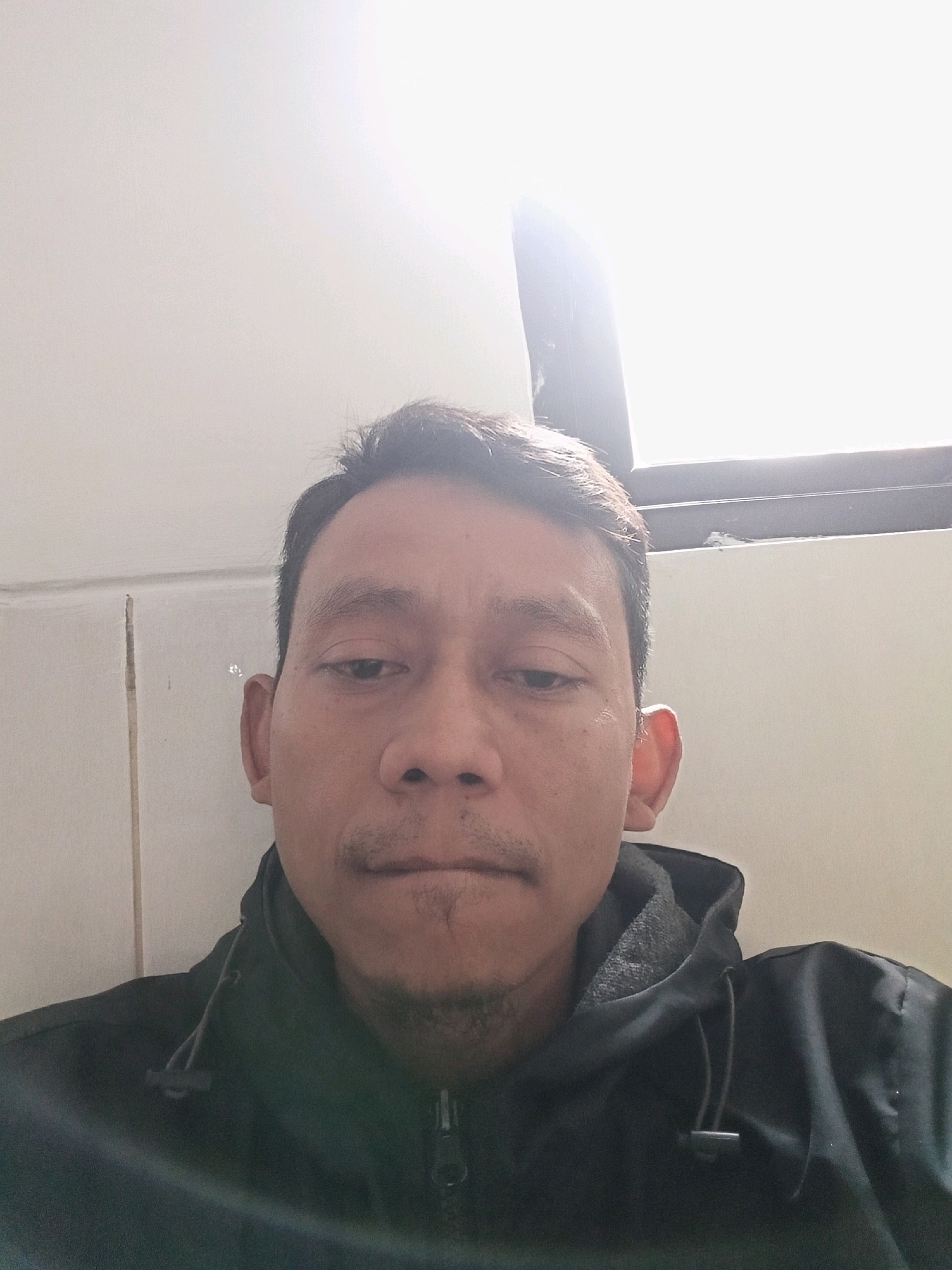 A man is captured in a head and shoulders selfie, looking directly at the camera. He has dark hair, styled short and swept back, and a light mustache and goatee. His complexion is tan, and he appears to be in his late 20s or early 30s. He is wearing a black hooded jacket with a zipper in the front. The background is a plain, off-white wall, possibly with some subtle texture or seams visible, and a bright light source is visible in the upper right corner, suggesting a window or strong overhead lighting. The setting appears to be indoors, given the wall and controlled lighting. The man is the sole subject of the image.