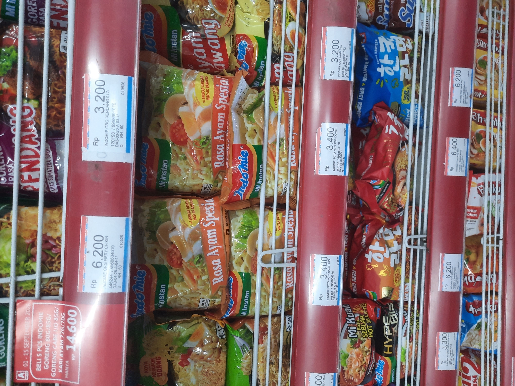 The image depicts a retail shelf display of instant noodles in a supermarket. The products are arranged in rows on white wire shelves, illuminated by overhead lighting. Several brands and flavors of instant noodles are visible, including Indomie. Price tags are displayed in front of some of the product stacks. One prominent display on the left shows a promotional offer for Indomie Goreng and Indomie Goreng Kari Ayam, priced at RP 14,600 for 5 pieces. Other visible prices include RP 6,200, RP 3,200, and RP 3,400 for various Indomie products. The background shows more shelves with packaged goods, and the overall environment suggests a well-stocked retail setting. The specific location in North Jakarta, Indonesia, cannot be determined from the image alone.