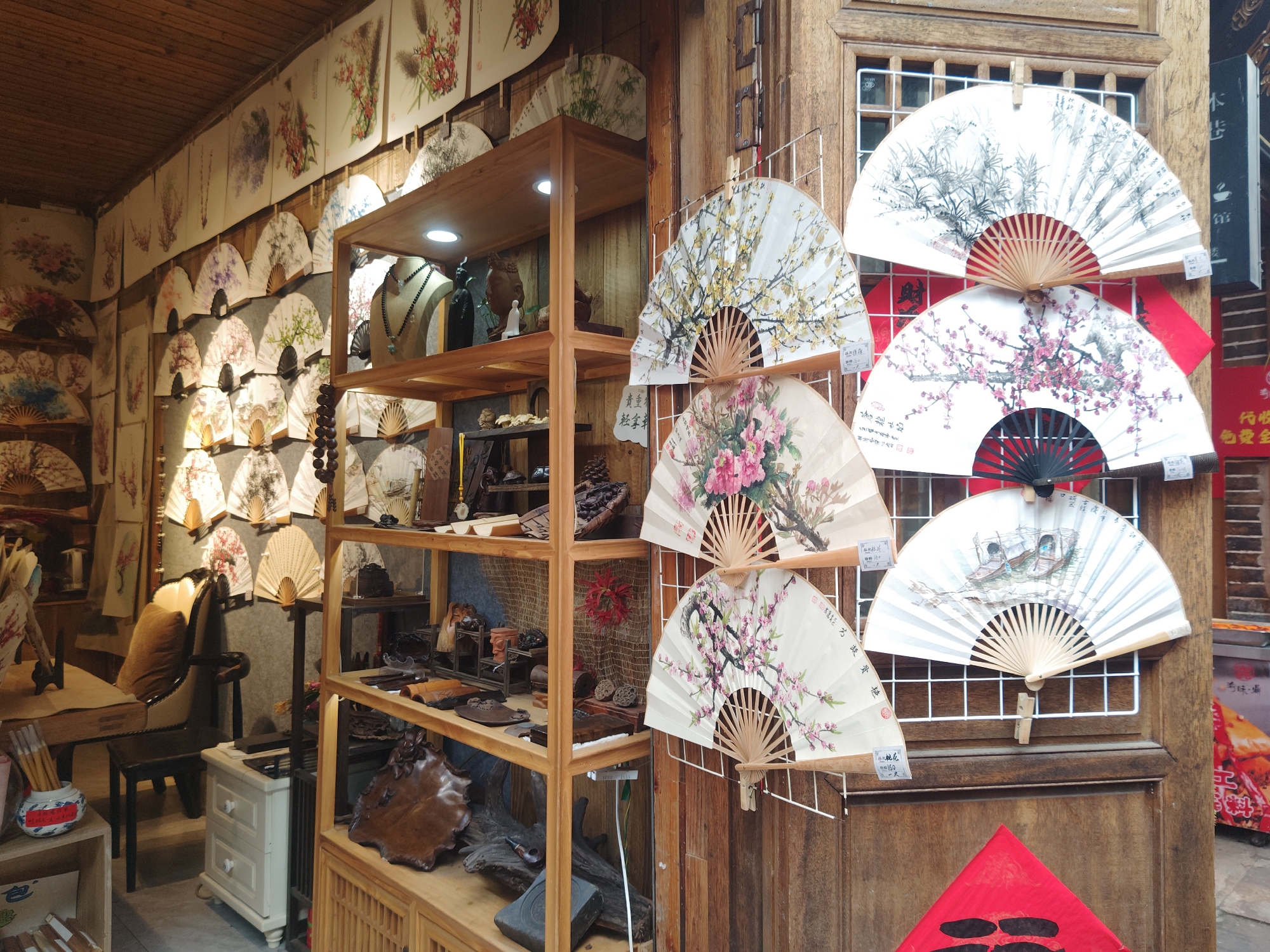 The interior of a shop in Shapingba District, China, features a wall lined with framed artwork and paintings, primarily of floral and natural scenes. A wooden shelving unit is filled with decorative items and crafts, including jewelry and carvings. Adjacent to the shelves, a display of various hand fans with different floral patterns is mounted on a wooden wall. In the background, a chair and table setting are visible, along with a small cabinet. The store's overall aesthetic is that of a traditional Chinese craft shop.