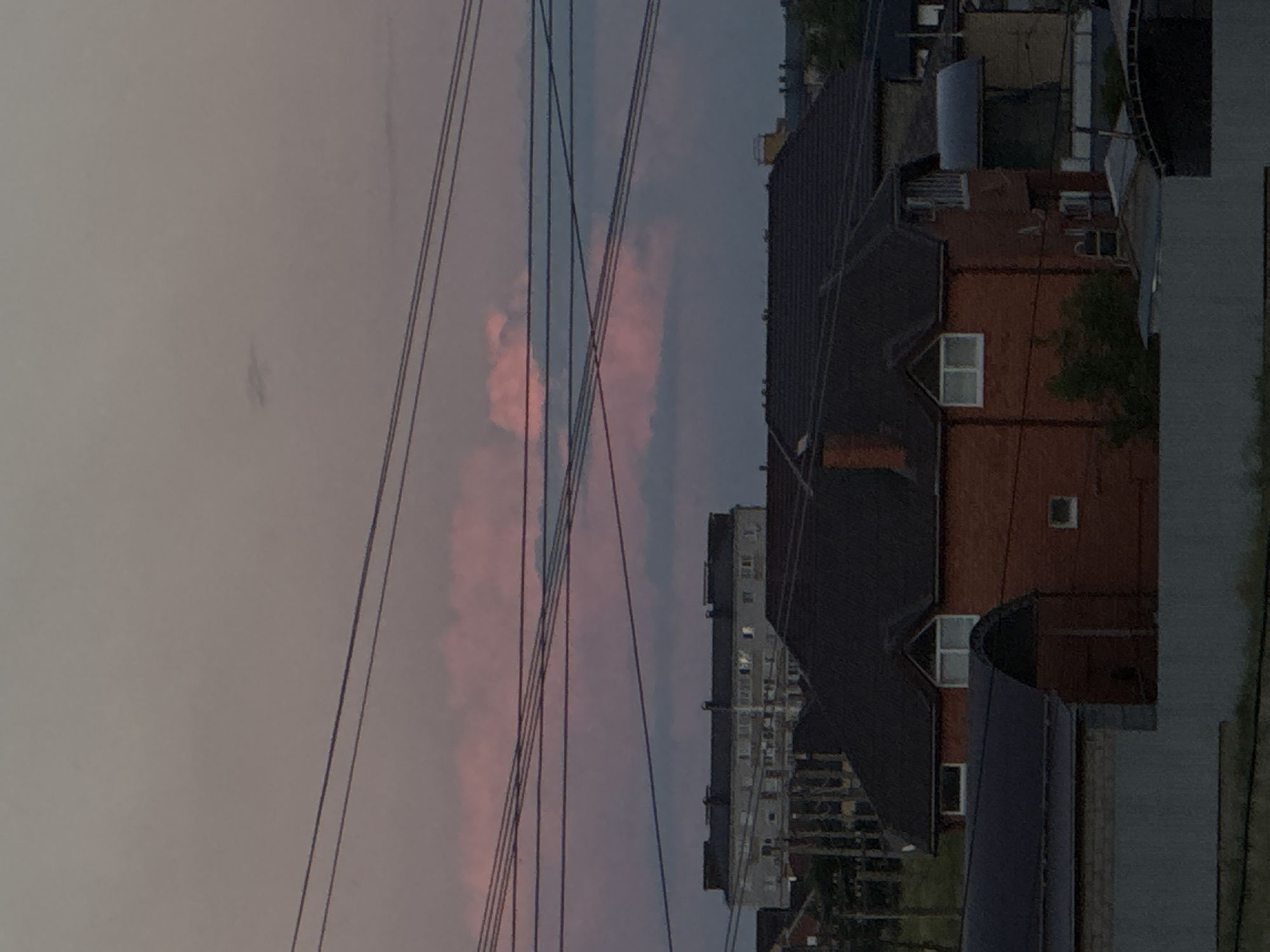 The image depicts a view of urban buildings against a twilight sky. The sky is a blend of gray and pink hues, suggesting either sunset or sunrise. Several dark, parallel wires run diagonally across the frame from the left to the right, obscuring parts of the scene behind them.
In the foreground, on the right side of the image, a building with a dark roof and red brick walls is prominent. It has several windows, some of which are visible and appear to be illuminated. Further into the background, a taller, multi-story building with numerous windows is visible. The overall scene suggests an urban environment observed from an elevated position, possibly a balcony or a higher floor of a building. The time of day is clearly dusk or dawn, indicated by the soft, colored light in the sky. There are no people or discernible text in the image. The mood is calm and somewhat muted, typical of the transition between day and night.