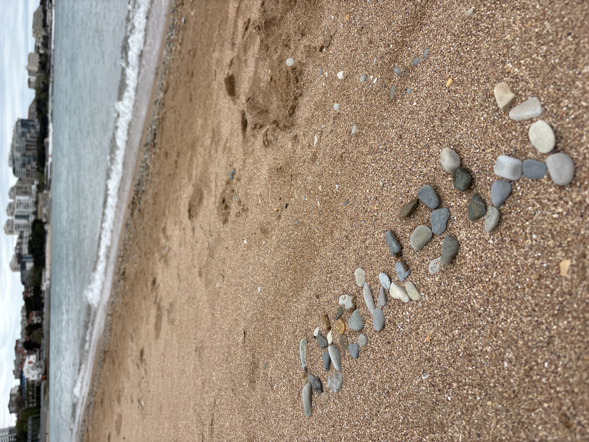 The image captures a beach scene in Gelendzhik, Russia, with a sandy beach leading to a body of water bordered by buildings in the distance. On the beach, small rocks are arranged in a line or arrow-like formation. The sand appears to be medium-grained and damp, with some footprint impressions. The water has gentle waves approaching the shore. The buildings visible across the water are multi-story structures typical of a seaside urban area. The sky overhead is overcast, suggesting a cloudy day.