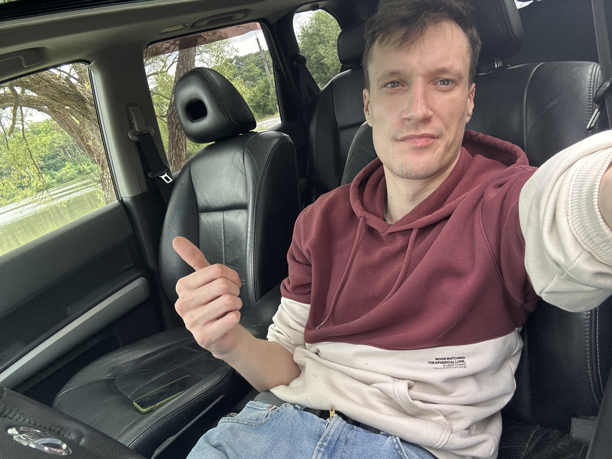 A selfie taken from the driver's seat of a car shows a young man in a maroon and cream hoodie and blue jeans. He is giving a thumbs-up gesture. The car's interior is visible, with black leather seats and a phone lying on the passenger seat. Outside the car, a body of water and lush green trees are visible, suggesting a rural or park-like setting. The scene is not in Makiv, Ukraine.
