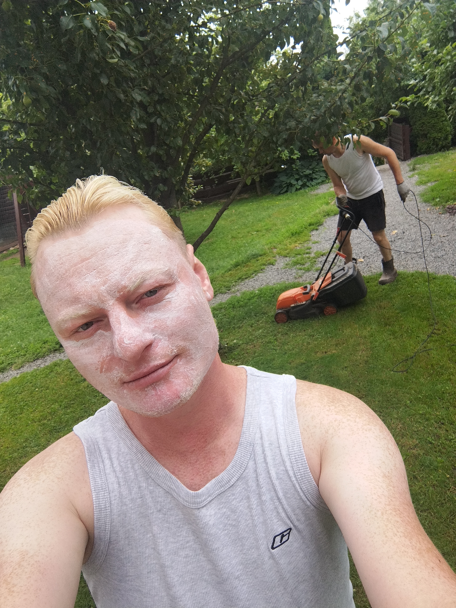 In a backyard setting, a man with fair skin and blonde hair, covered in a white substance on his face, is taking a selfie. He is wearing a grey tank top. In the background, another man is pushing an orange and black lawnmower across a grassy area. The environment appears to be a residential yard with trees and foliage. No specific location in Mahilyow, Belarus can be identified from the image.