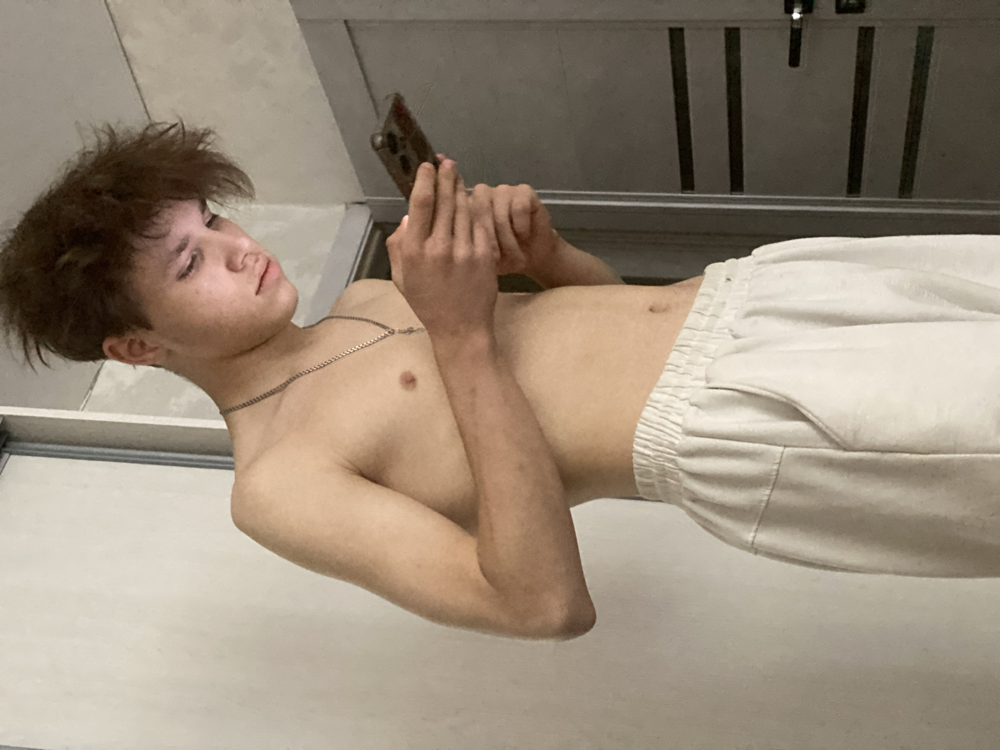 A young male, appearing to be a teenager or young adult with light skin and messy, medium-brown hair, is captured indoors. He is lying down, positioned with his head to the left and body extending to the right, fully engaged with a smartphone held in both hands. A thin silver chain necklace adorns his neck, and he is shirtless, wearing only light-colored (likely white or off-white) drawstring pants or shorts. His gaze is directed intently at the phone screen, and his expression appears calm and focused.
The immediate background behind his head and shoulders is a plain, light-colored surface, possibly a wall or bedding. Beneath him, there is also a light-colored surface. To the right of the frame, behind his lower body, a darker, textured background featuring vertical slats or panels suggests an interior fixture, such as a closet door or room divider. The lighting is soft and even, indicating an indoor setting, and no specific time of day is discernible. There is no visible text in the image. This scene is located in Sochi, Russia.