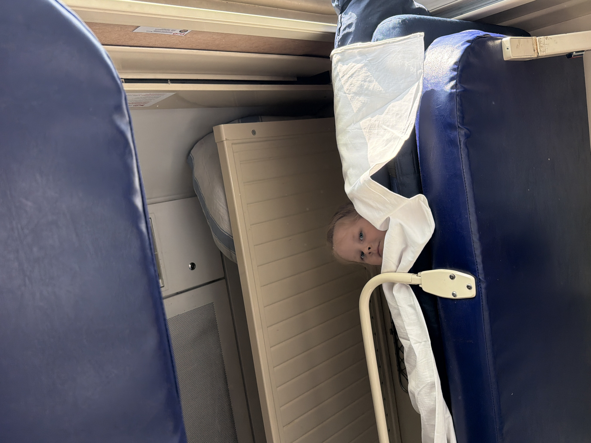 A close-up shot, taken inside a compartment of a train, likely in Russia. A fair-skinned child with light blonde hair and blue eyes, partially covered by a white sheet, peeks out from between a beige fold-down bed and a blue padded seat. The seat's metal armrest is visible. The wall is a light grey, and the overall environment is confined and utilitarian. Karamysh is not visibly discernible from the image.