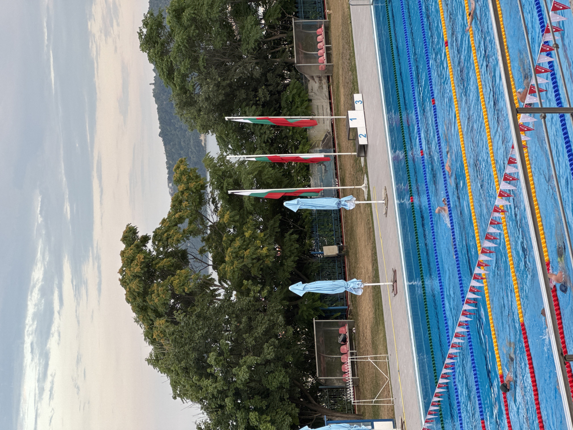 An outdoor swimming pool complex is depicted in Varna, Bulgaria, likely during a swimming competition given the presence of swimmers in lanes and a podium. The pool is large, with multiple lanes marked by buoys in red, yellow, and blue. Several swimmers are visible in the water, actively swimming. Flags of Bulgaria are flying on flagpoles. Blue and white umbrellas are positioned near the edge of the pool. In the background, there are bleachers with red seats and a wooded area with lush green trees. The sky is overcast with clouds. The image captures a moment of athletic activity within a recreational or sporting venue.