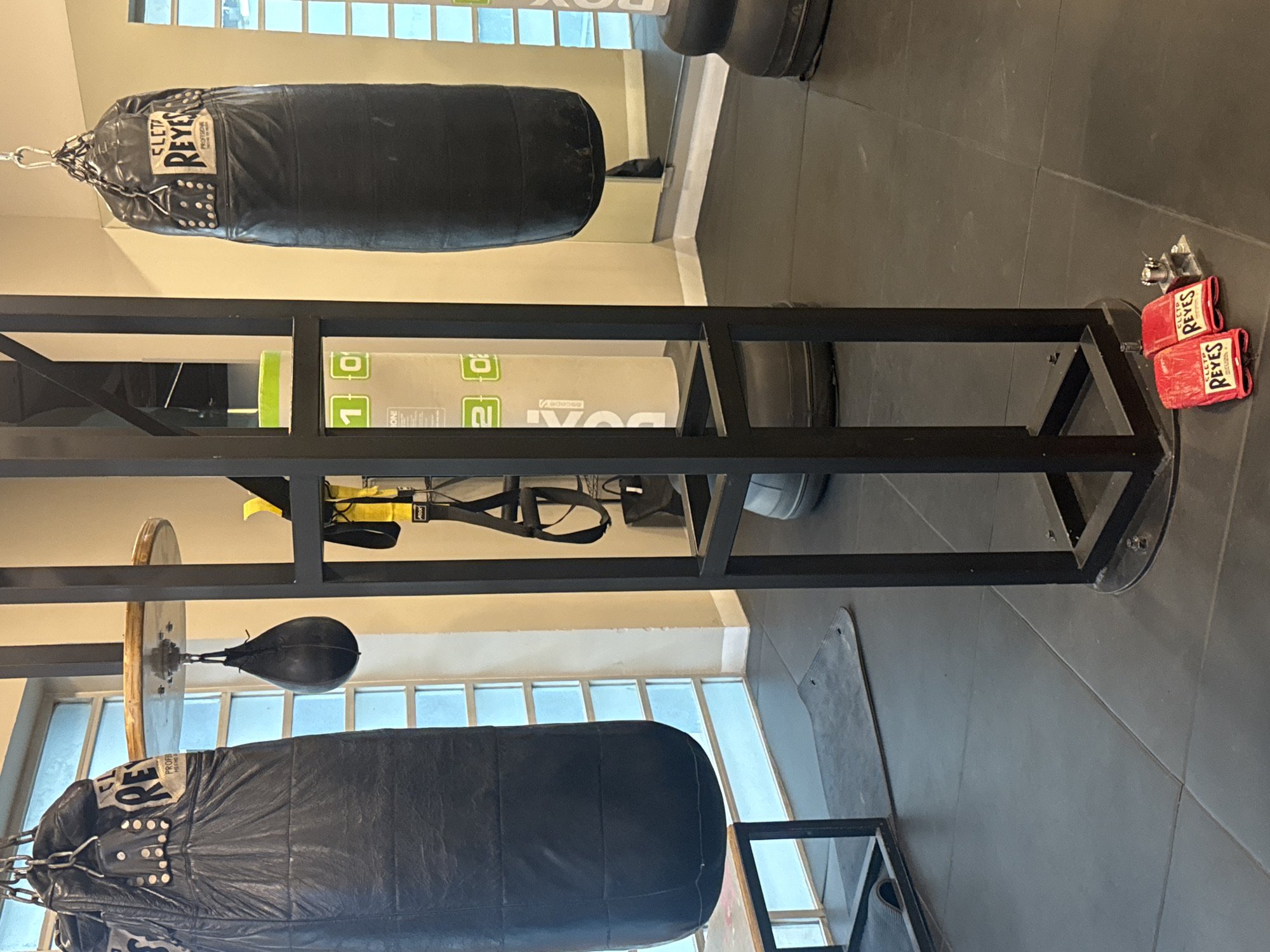 This media file presents an indoor gym scene, rotated 90 degrees counter-clockwise. The setting appears to be a boxing or fitness training facility.
**Subjects:** The primary subjects are two black heavy punching bags and one black speed bag, suspended from a robust black metal frame structure. A pair of red boxing gloves lies on the dark gray tiled floor. In the background, reflections in a mirror reveal windows, a white mat or target, and other gym equipment. TRX suspension trainers are also visible hanging from the frame.
**Activity or Event:** No individuals are present, so no specific activity is currently in progress. However, the presence of boxing bags, a speed bag, and boxing gloves clearly indicates that the area is used for boxing or general fitness training.
**Notable Details:** The image is well-lit, suggesting daytime, with light entering through the windows. The large black metal frame serves as a central support for the punching bags, which appear to be made of leather or a similar durable material. One heavy bag hangs horizontally in the upper left, while another is in the lower left, beneath a speed bag attached to a wooden circular platform. The red boxing gloves are positioned near one of the frame's support legs on the right. The floor is covered with dark gray tiles.
**Visible Text:** The brand "CLETO REYES" is clearly visible on both the heavy punching bags and the red boxing gloves. Additionally, the words "PROFESSIONAL" and "HECHO EN MEXICO" are legible on the punching bags. In the background reflection, text including "BOX," "ESCAPE," and numbers "1" and "2" can be seen on a white object. The "TRX" logo is visible on the black suspension straps.