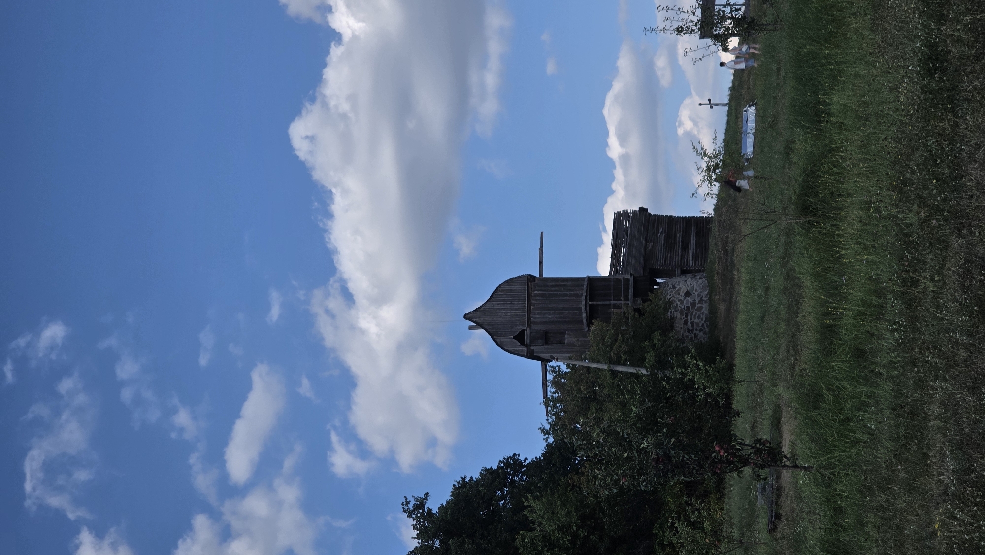 The image captures an old wooden windmill situated on a grassy hill under a partly cloudy blue sky. The windmill, appears to be a historical structure, likely part of an open-air museum or preserved rural site. In the background, a few people are visible on the hill, one of whom is near a reflective object, possibly a picnic blanket or a tarp. A cross is also visible on the hill in the middle ground, suggesting a possible religious or historical significance of the location. The foreground is dominated by green grass and some young trees. The overall setting evokes a rural or park-like atmosphere. This scene is consistent with locations such as the Pirogovo Open Air Museum in Kyiv, Ukraine, which features historical wooden architecture and is set in a natural landscape.
