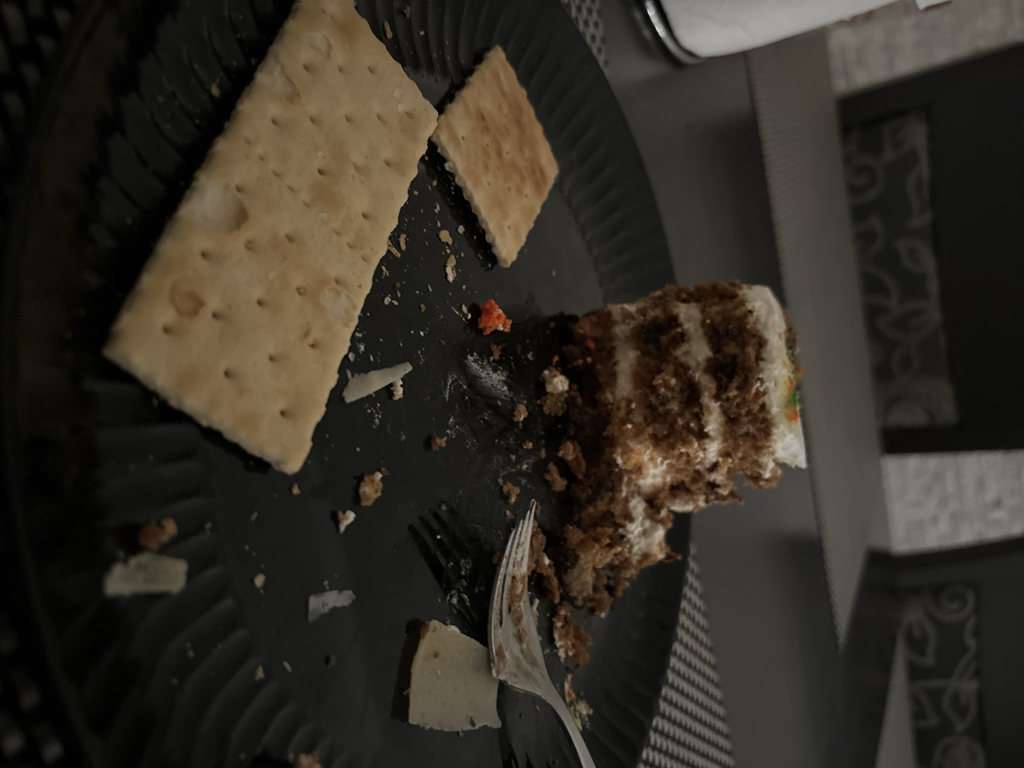 The image shows a close-up, overhead view of a black plate with leftover food, likely a dessert. Two rectangular crackers are prominently displayed on the left side of the plate, with one significantly larger than the other. A slice of cake, appearing to be carrot cake with cream cheese frosting, is partially consumed on the right side. Crumbs and small pieces of the cake are scattered across the plate, along with a few small, white pieces that could be cheese or more frosting. A silver fork is positioned near the cake, with its tines partially embedded in the dessert. There are also a few small orange specks on the plate, possibly sprinkles. The background is dark and out of focus, but suggests an indoor setting with patterned surfaces and furniture. The lighting is dim, creating a somber mood, and it is not possible to determine the time of day or weather. There is no visible text in the image. The scene appears to be a casual, domestic setting in Budva, Montenegro, where a meal or snack has recently concluded.