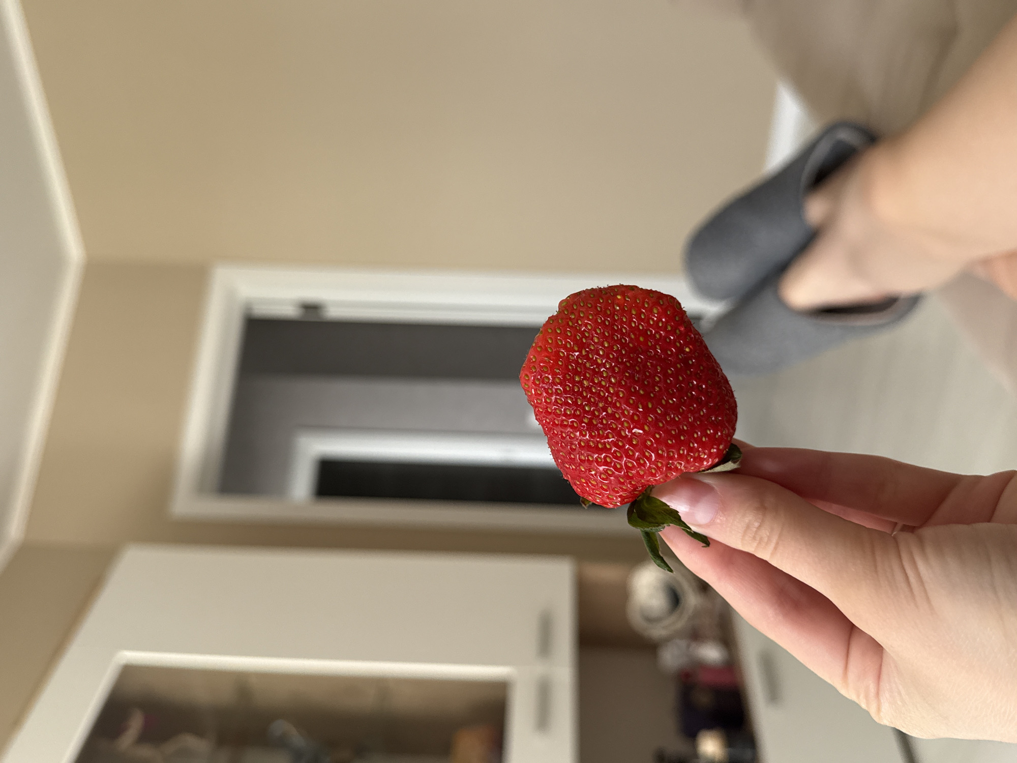 The image displays a prominent, large, bright red strawberry held by a person's hand. The strawberry has a textured surface with many visible small, yellowish seeds and retains its green stem and calyx, which the hand is grasping. The hand has light-toned skin, and clear, manicured fingernails are visible on the thumb and index finger.
The background suggests an indoor setting, likely a home. On the left side, a light beige wall meets a white wall or trim. In the mid-ground, a white-framed doorway leads into a darker room or hallway, providing depth. Below the doorway, parts of white cabinets or furniture with a reflective or clear-fronted section are visible. In the upper right corner, another hand or foot is partially seen, holding or wearing a grey, soft-looking slipper or shoe. The background elements are largely out of focus, drawing attention to the strawberry in the foreground. The lighting appears to be ambient indoor light. No text is visible in the image.
