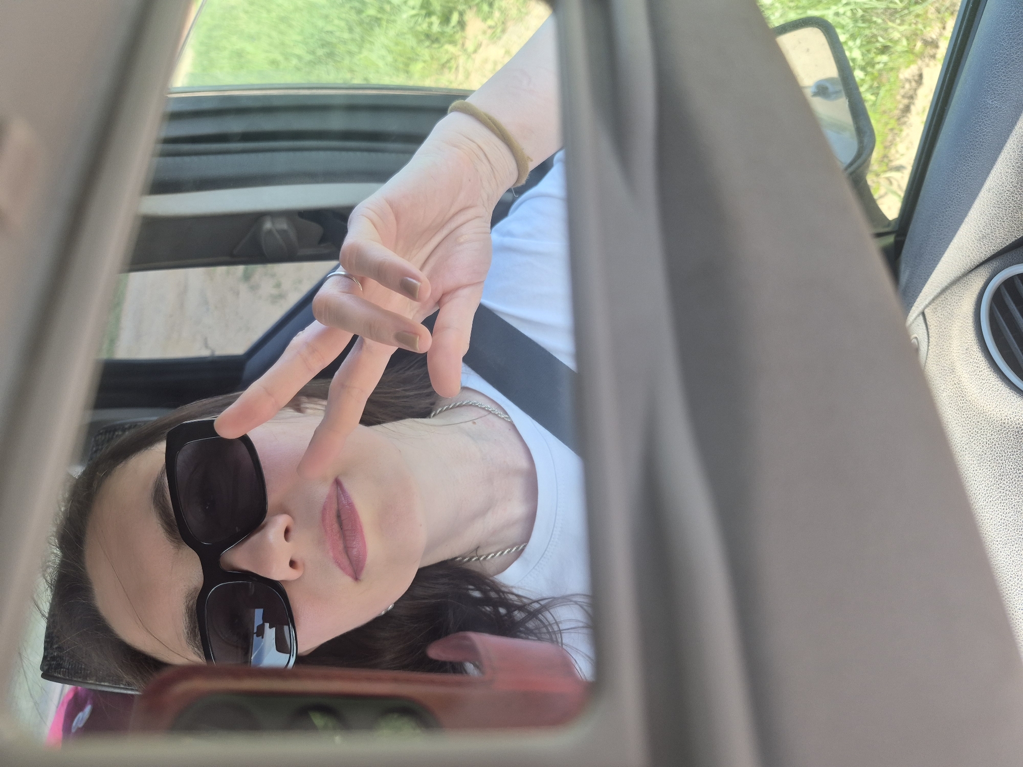 A reflection in a car's sun visor mirror shows a person wearing sunglasses and making a peace sign. The person has brown hair and is wearing a white shirt with a seatbelt. The car's interior and side mirror are visible. Beyond the car window, a road and greenery suggest a rural or semi-rural location.