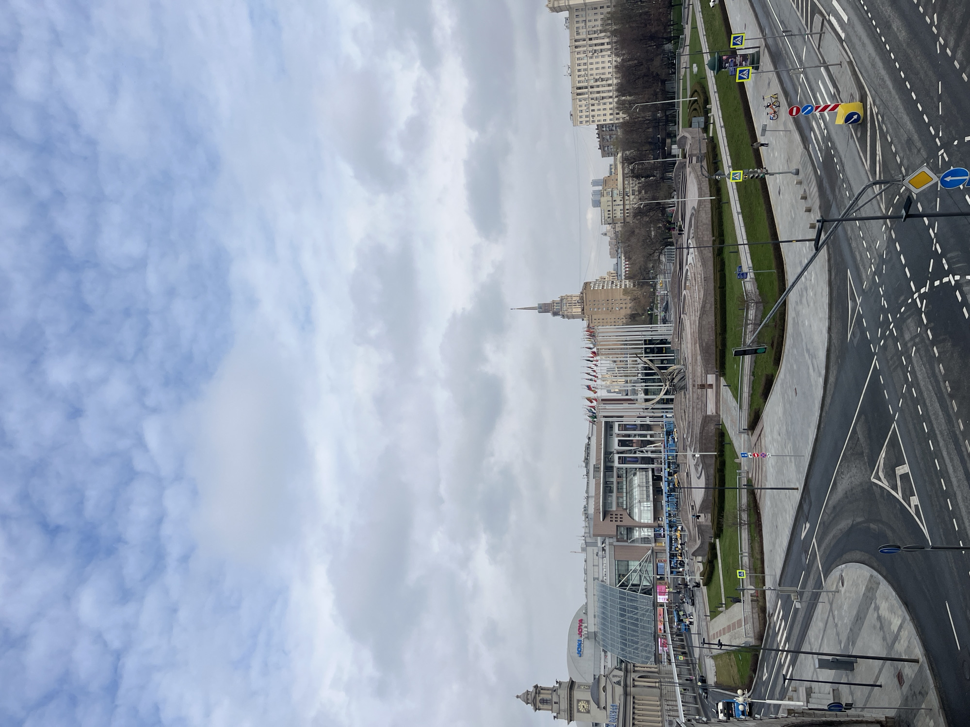 The image shows an elevated view of Moscow, Russia, on a cloudy day. Prominent landmarks include the Hotel Leningradskaya, one of the Seven Sisters skyscrapers, and the building housing the Tsvetnoy Central Market. A multi-lane road divides the foreground, marked with numerous white dashed lines and signage. Pedestrian walkways lined with manicured greenery parallel the road. A few people are visible along the walkways. Flags are seen near the Tsvetnoy Central Market, and various street signs are also present. The sky is mostly overcast.
