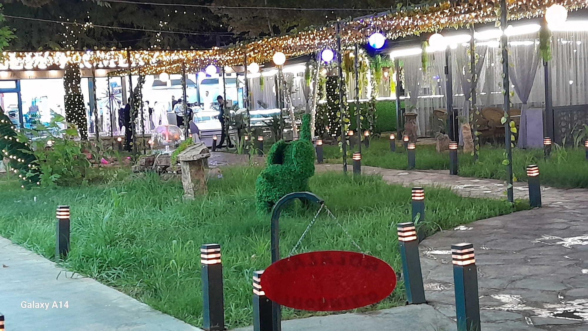 The image depicts an outdoor seating area decorated with string lights and greenery, likely a restaurant or cafe, located in Karshi, Uzbekistan. In the foreground, a red oval sign hangs from a metal stand, its surface obscured. The ground is paved with stone and surrounded by lush green grass. Several black bollard lights with illuminated tops are strategically placed along the path. In the background, a sculpted green bush resembling a sitting figure is visible. Beyond that, a building with large windows is adorned with numerous fairy lights, suggesting an evening setting. People are faintly visible inside the building and in the immediate outdoor area. The overall atmosphere suggests a well-lit, inviting outdoor space.