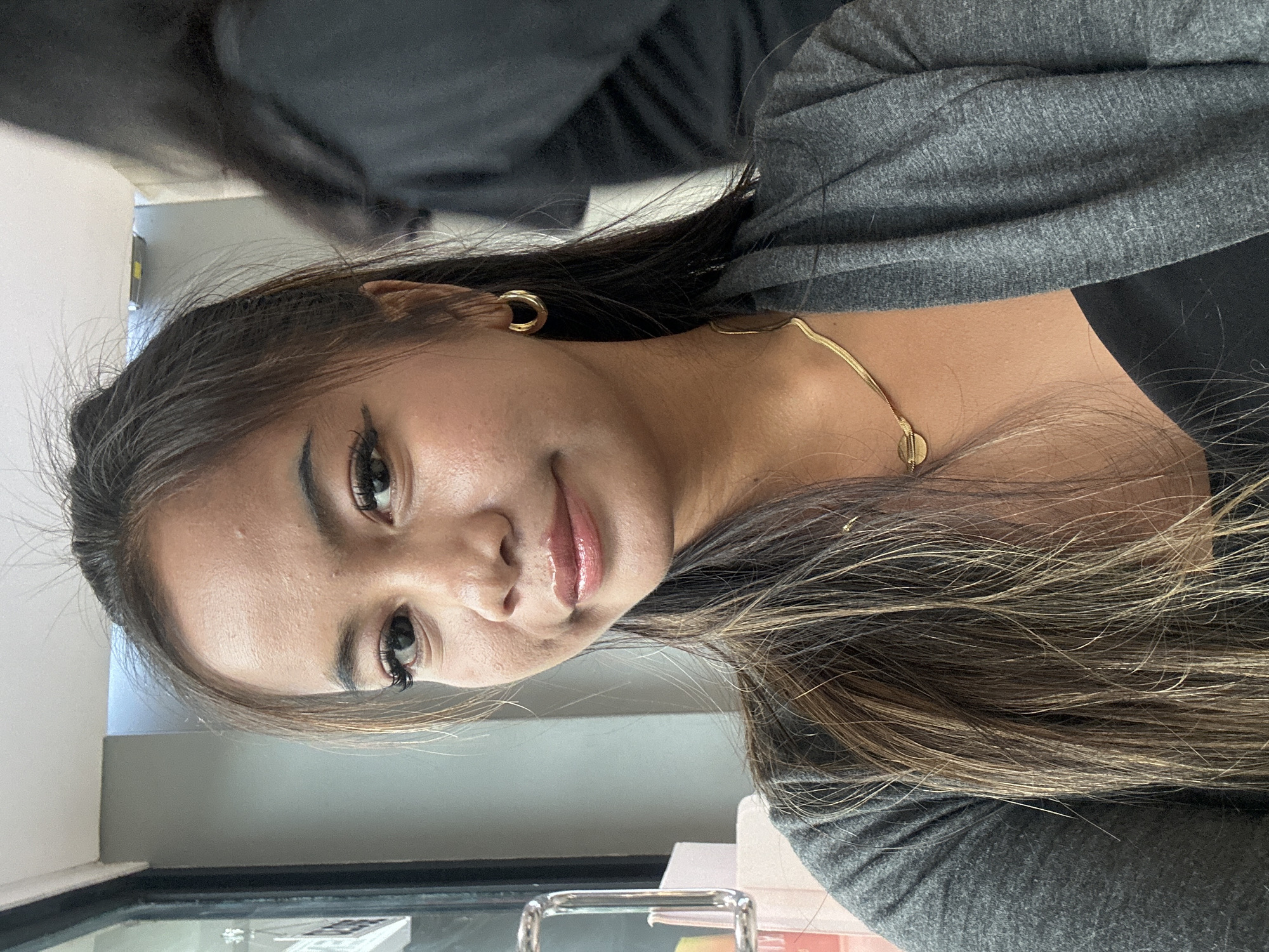 A woman is depicted in a close-up shot, facing the camera with a slight smile. She has long, dark hair with light brown highlights, gold hoop earrings, and a gold necklace. She is wearing a gray cardigan over a black top. The background appears to be an indoor setting, possibly an office or classroom, with a window and various items visible on a desk. There is no specific indication in the image that points to a location in North Jakarta, Indonesia.