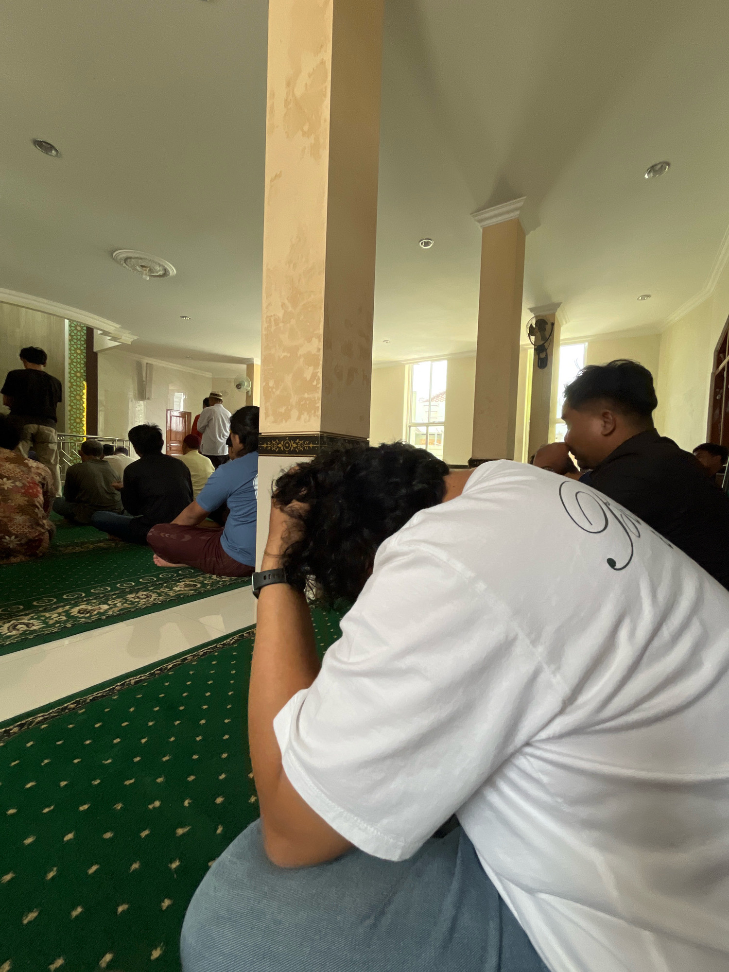 The image depicts a gathering of men seated on a green carpet with a repetitive pattern inside a mosque. The space is illuminated by natural light filtering through windows and overhead lighting. In the foreground, a man wearing a white t-shirt and blue jeans is seated, with his head bowed and hands near his face. Behind him, other men are visible, some facing forward and others to the side. One man in a black shirt is seated to the right of the frame. Pillars with textured surfaces are present within the mosque, dividing the space. The overall atmosphere appears to be one of prayer or quiet contemplation. The location is identified as Gentan, Indonesia.