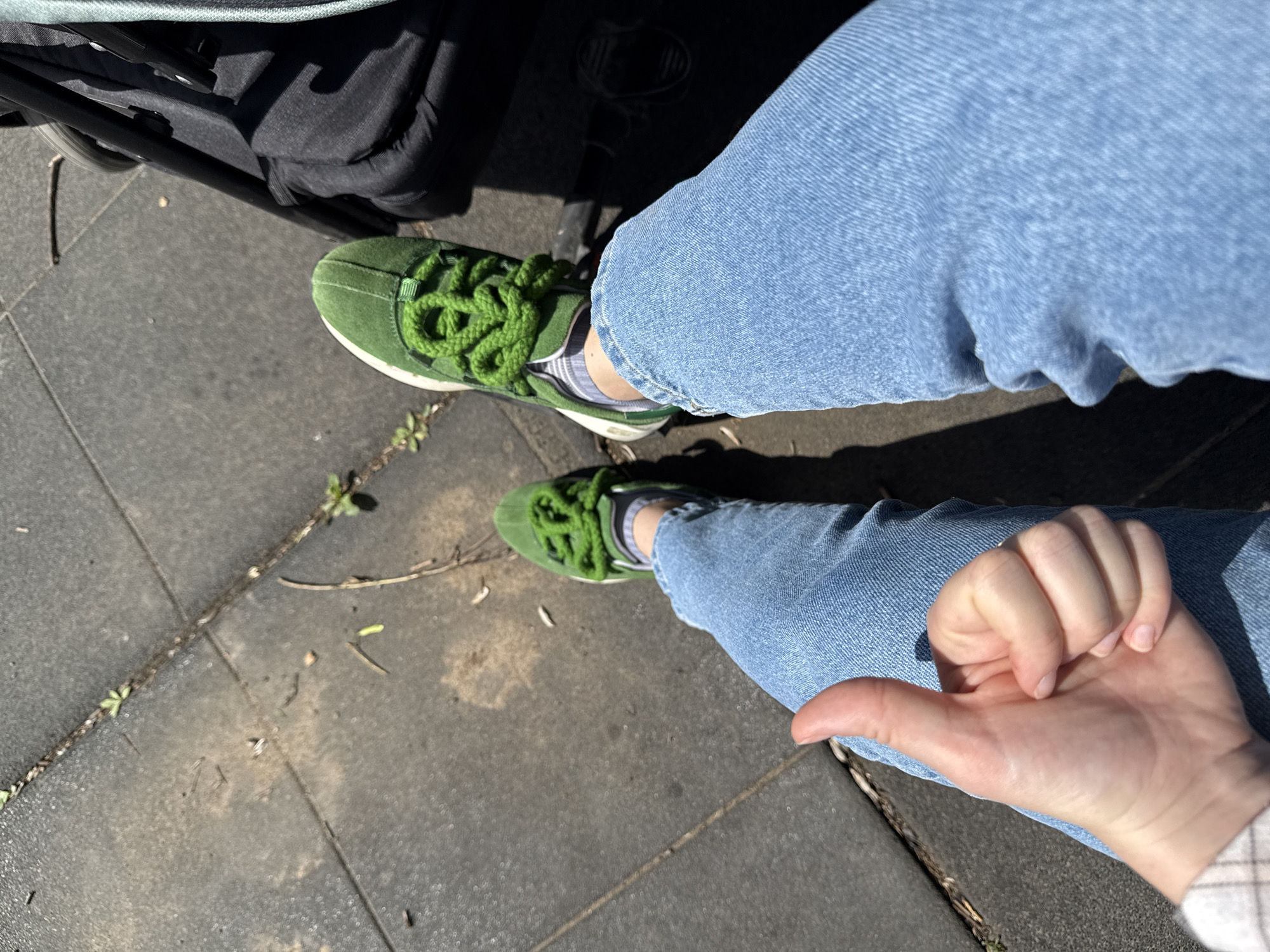 A first-person perspective, overhead shot shows a person sitting on a grey tiled surface, with their legs extended. The person is wearing light blue jeans and a pair of bright green sneakers. The sneakers have thick, bright green laces that are loosely tied. The person's right hand is in the foreground, giving a thumbs-up gesture. To the right of the person's legs, a black stroller with a grey canopy is partially visible. The ground appears to be outdoors, with sunlight casting shadows. The environment is likely a park or a public walkway in a city, given the tiled ground and the stroller.
