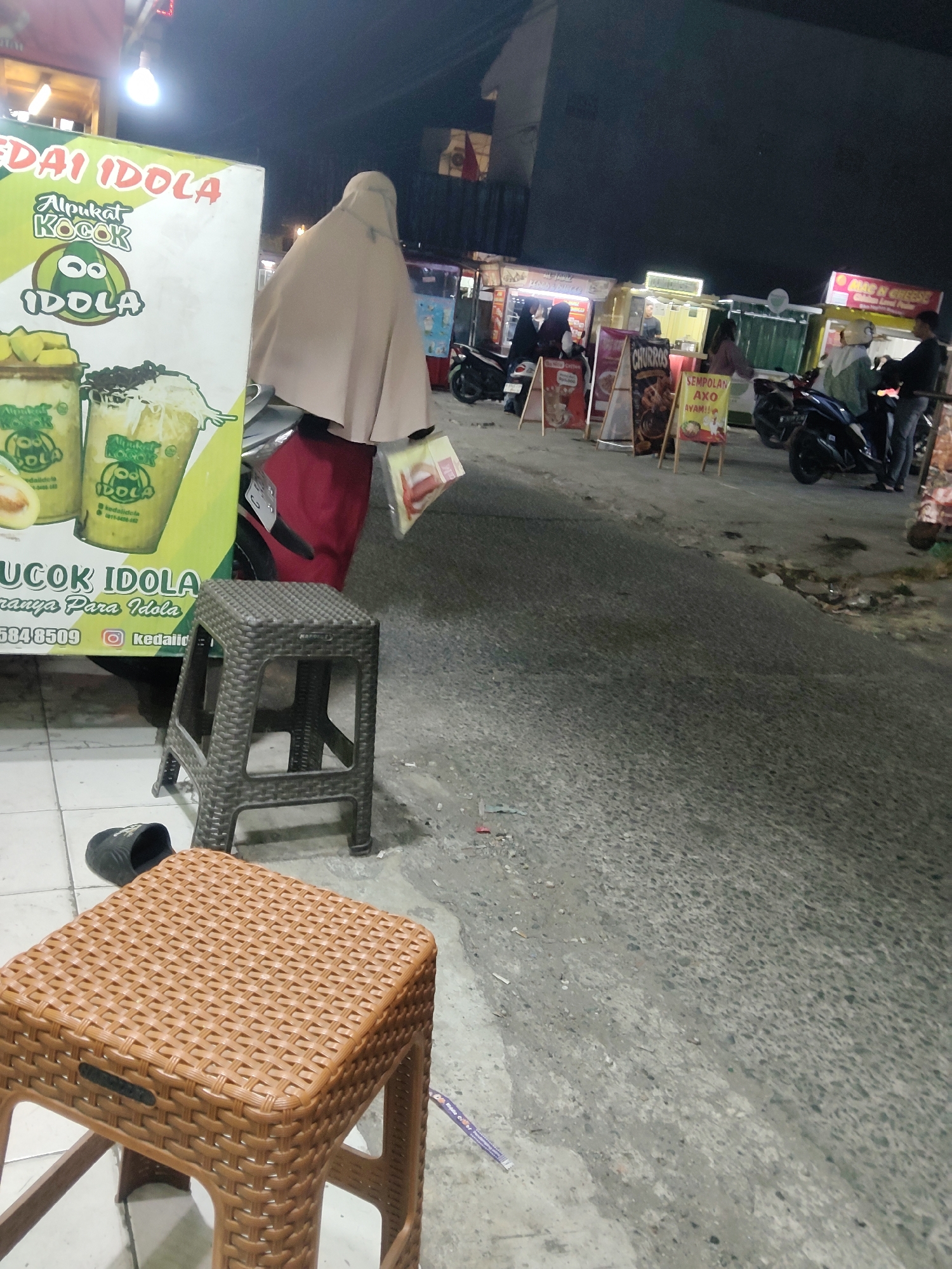 The image depicts a nighttime scene at "KEDAI IDOLA" in Mekarmukti, Indonesia. A large green sign for "KEDAI IDOLA" advertises "Alpukat Kocok" (avocado shake) with imagery of the drink. In front of the sign, two plastic stools, one dark brown and one woven brown, are placed on the ground. A person wearing a light brown hijab and a red garment is partially visible behind the sign. In the background, several food stalls line a street, with motorcycles parked nearby. People are seen interacting at the stalls, and promotional signs for items like "CHURROS" and "SEMPOLAN AXO AYAM" are displayed. The environment appears to be an outdoor street market or a roadside food vendor area.