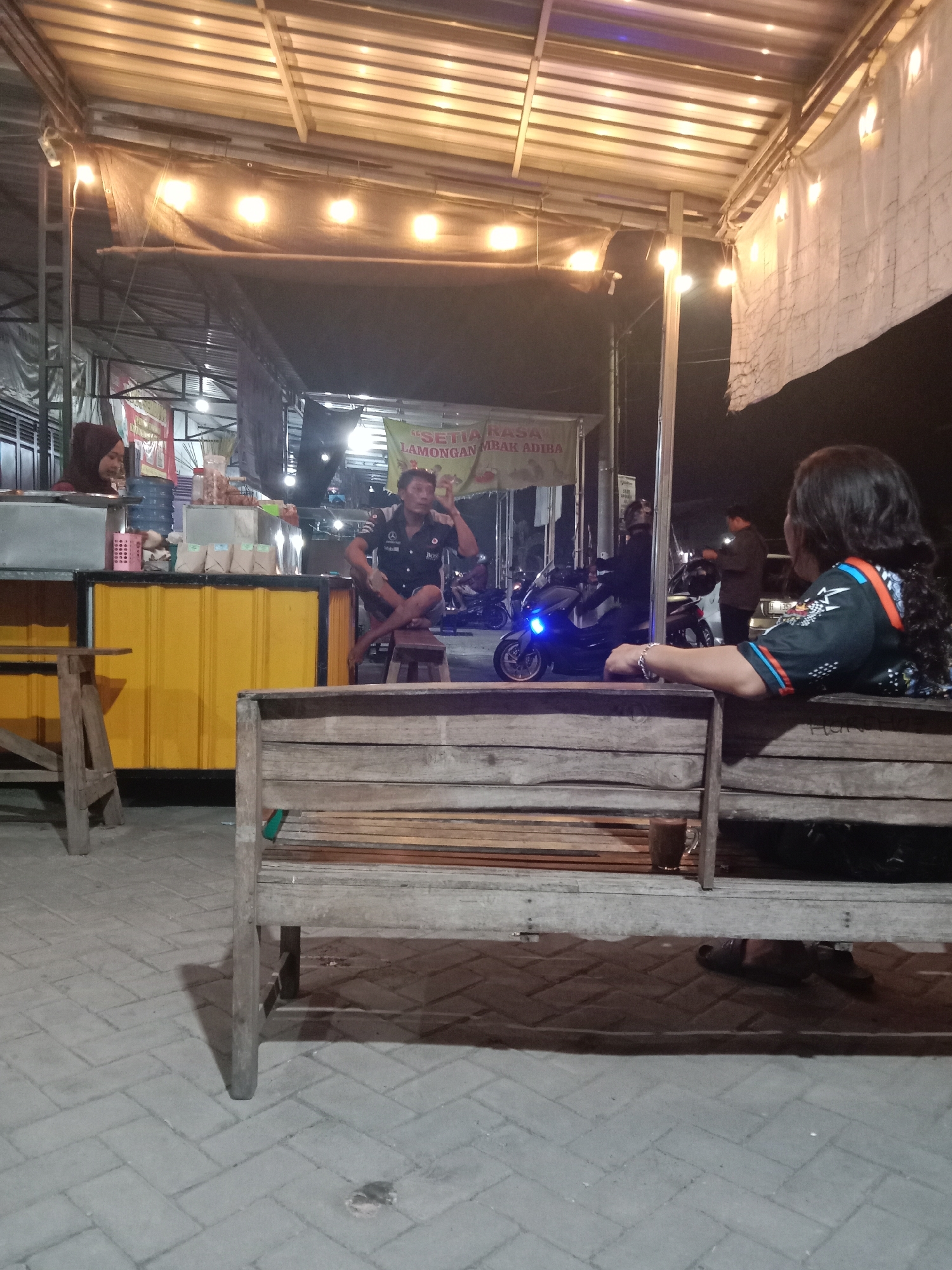 This is a night-time, medium-shot photograph taken at a street food stall or outdoor eatery in Cangkring, Indonesia. The scene is illuminated by overhead string lights. In the foreground, a wooden bench is visible, with a glass of dark liquid, likely a beverage, placed on it. A person with long, dark, curly hair, wearing a black shirt with colorful trim, is seated on the bench facing away from the camera.
In the middle ground, a man in a dark uniform with patches, including a Mercedes-Benz logo and the word "Mobil," is sitting on a stool and appears to be speaking or gesturing, with his right hand near his head. Behind him, a yellow counter is visible, with various items on display, including bottles and packets. To the left of the man, a woman wearing a hijab is standing behind the counter. Further back, several motorcycles are parked, one of which has its headlights on. There are also banners with Indonesian text visible, one appearing to read "SETIA LAMONGAN MBAK ADIBA". The ground is paved with interlocking grey bricks. The overall atmosphere suggests a casual, open-air dining or gathering spot.