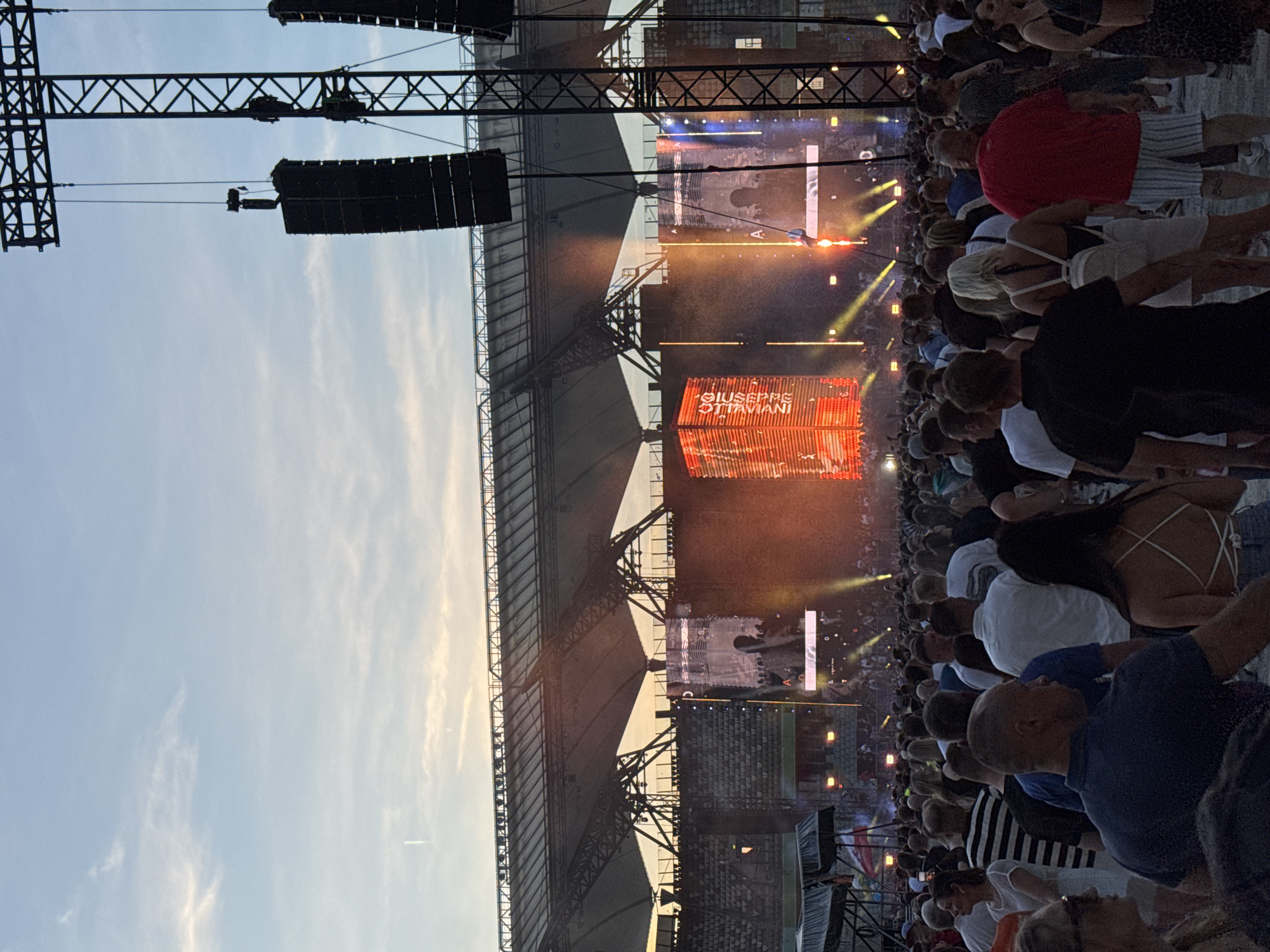 The image depicts a large outdoor concert at PGE Narodowy stadium in Warsaw, Poland. A dense crowd of spectators is visible, facing a brightly lit stage with large screens and sound equipment. The sky is clear with a hint of sunset colors. The stadium's roof structure is prominent in the background.