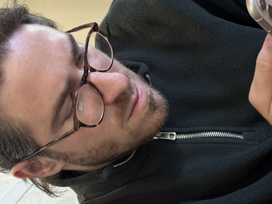 A close-up, eye-level image depicts an adult male, with dark hair pulled back and short facial hair, looking downwards. He wears tortoiseshell-patterned eyeglasses, exhibiting reflections on the lenses. The individual is dressed in a dark green or dark grey fleece pullover, featuring a partially unzipped silver-colored zipper extending down the front. His right hand is visible in the lower right portion of the frame, holding a light-colored smartphone, a section of which including part of its camera array is discernible. The background is out of focus, showing indistinct, light-colored areas consistent with an indoor setting. The image provides no verifiable visual data to confirm a location within Tutin Municipality, Serbia.