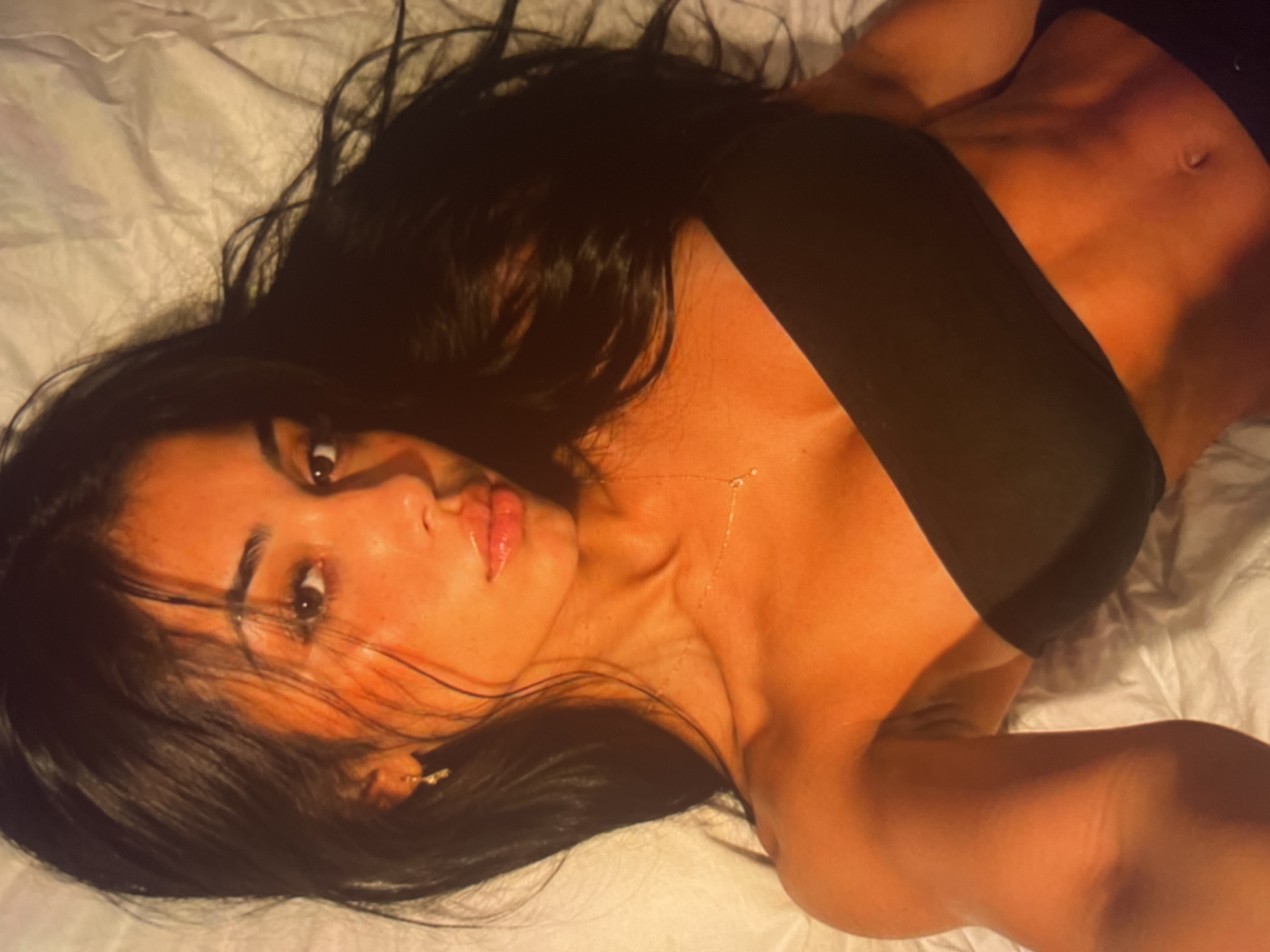 A young woman with long, dark hair lies on her back on a white, wrinkled surface that appears to be a bed. She is wearing a black bandeau top and black bottoms. Her head is tilted back, and her eyes are looking up and to her right. The lighting is warm and casts a golden hue over her skin, highlighting her facial features and toned midriff. A delicate gold necklace is visible around her neck. The image is a close-up shot, focusing on her upper body.