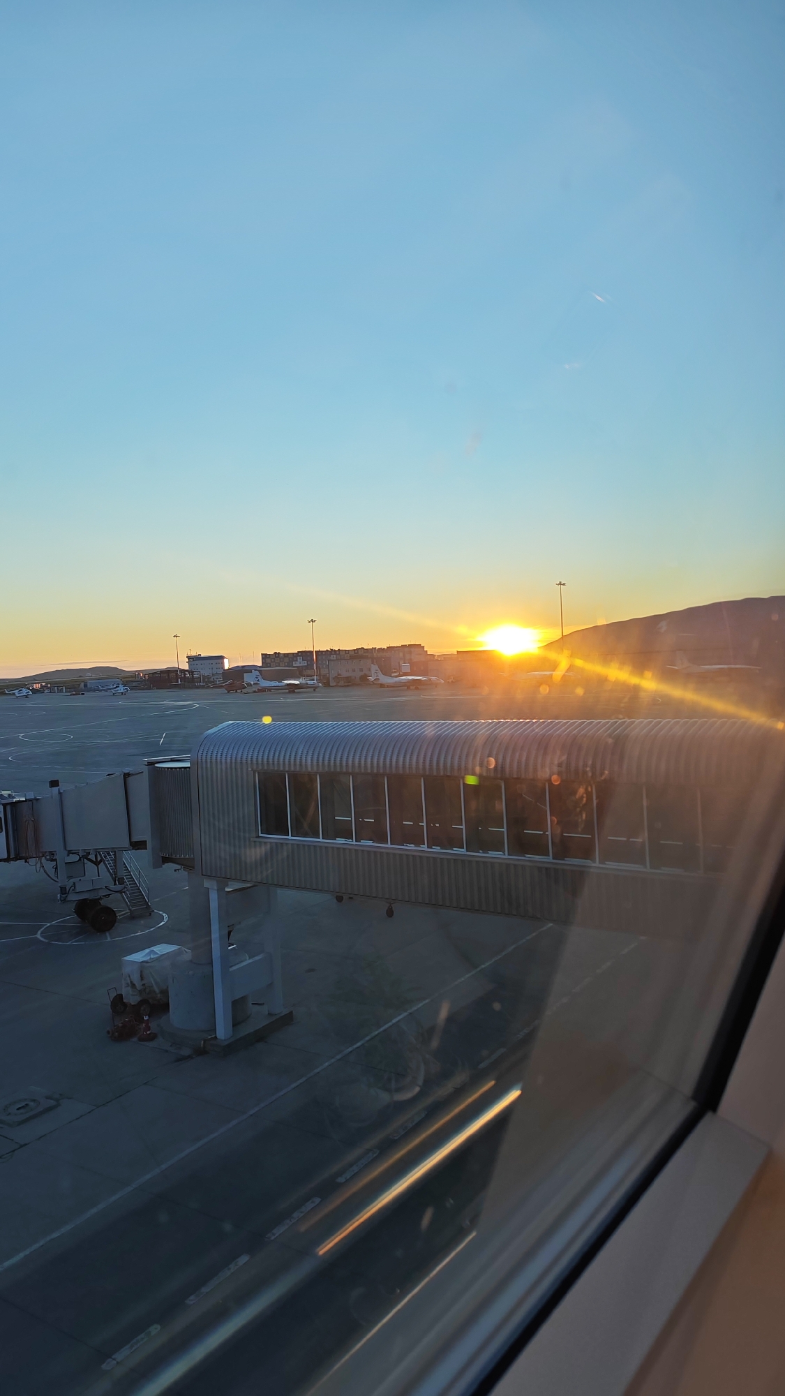 The image captures a sunrise at an airport in Ugolnye Kopi, Russia. The sun is low on the horizon, casting a warm, golden glow across the scene. An airport jet bridge, a covered walkway connecting an airport terminal to an aircraft, is prominently featured in the foreground, extending across the tarmac. Several aircraft are visible parked on the tarmac, along with airport infrastructure such as buildings and light poles. The sky is clear and a soft blue, transitioning to warmer hues near the horizon. The overall impression is one of a calm, early morning at an airport, bathed in the gentle light of the rising sun.
