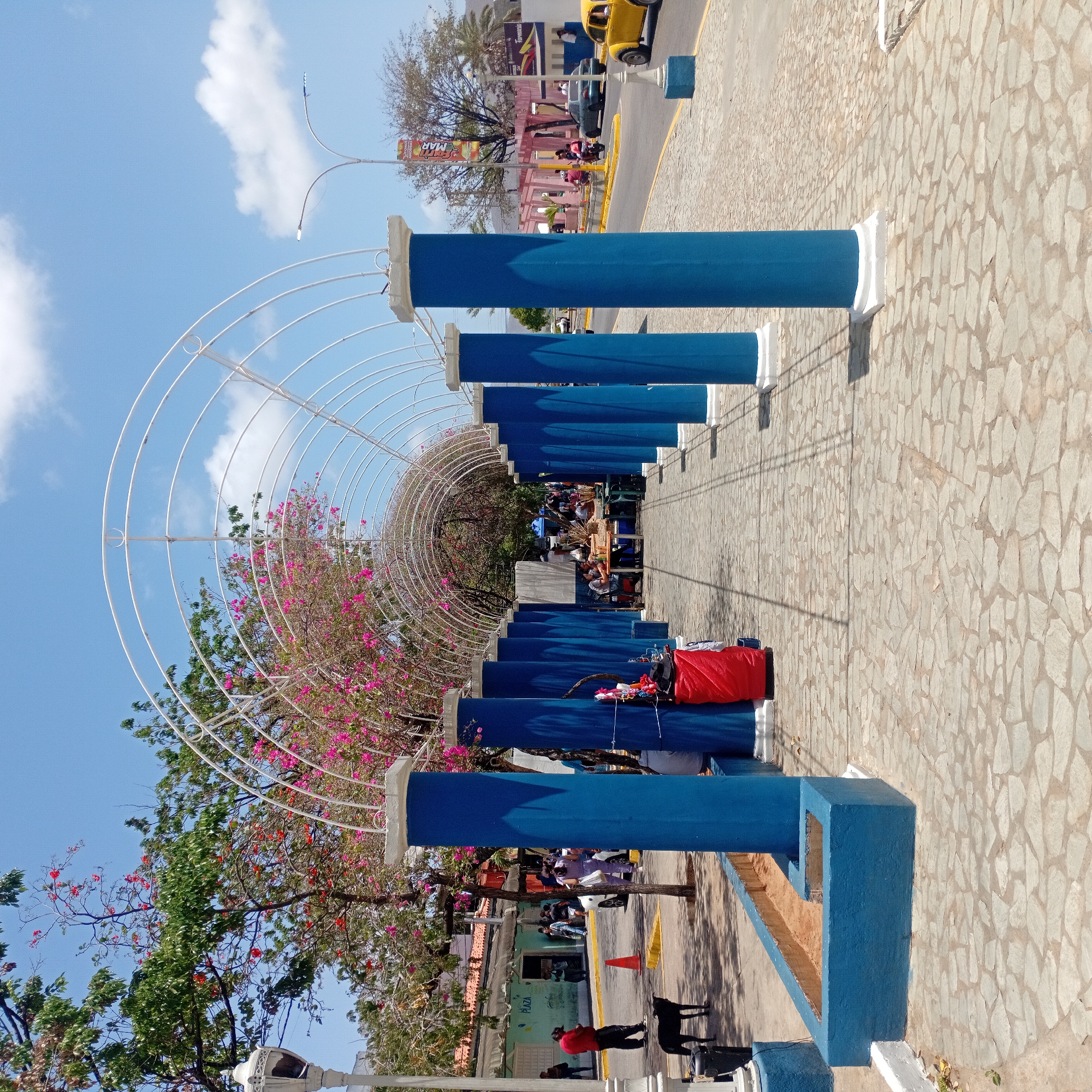 The image captures a walkway in an unidentified city, with a series of blue pillars supporting a white arched structure overhead. The walkway is paved with irregularly shaped stones. People are present along the walkway and adjacent to it, with a vendor displaying goods. A red bag sits atop one of the blue pillars. Trees with pink and green foliage intermingle with the arched structure. Beyond the walkway, there's a street with cars and various buildings. The sky is blue with scattered clouds.
