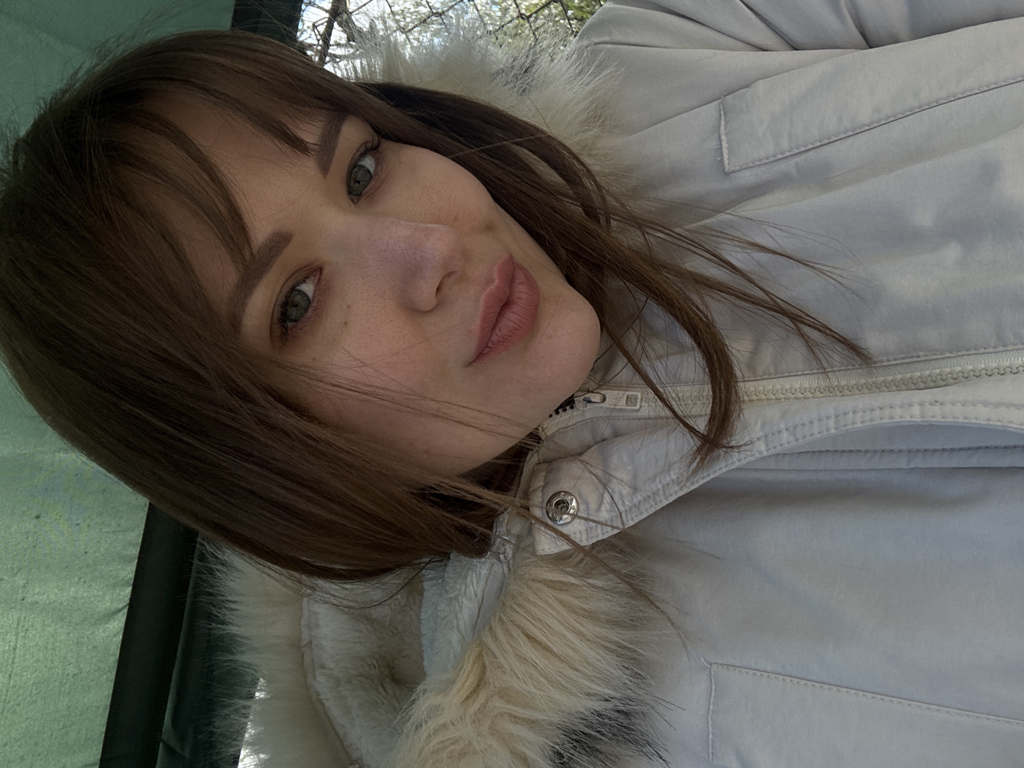 A woman with medium-length brown hair and bangs is wearing a light-colored jacket with a fur-trimmed hood. The jacket is zipped up to the collar. She is looking directly at the camera. The background shows glimpses of green foliage and branches, suggesting an outdoor setting. The exact location in the city of Chaikovsky, Russia, is not discernible from the image.