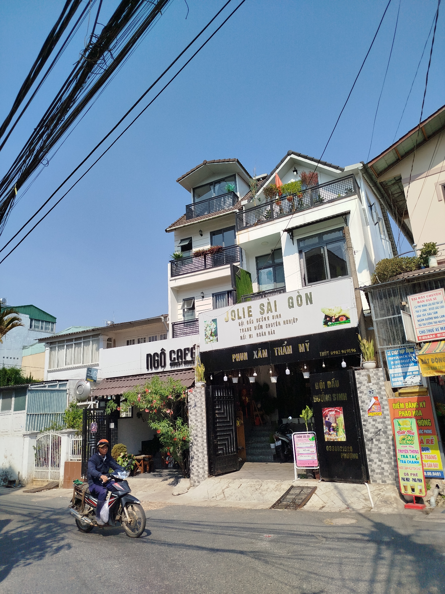 A street scene in Da Lat, Vietnam. A man rides a motorcycle on the road. Buildings are visible on the right side of the road, displaying various signs and advertisements. The building directly behind him is “Jolie Saigon” Beauty Center with the signs “Ngo Cafe.” Electrical wires run overhead against a clear blue sky. The environment appears to be a bustling area with multiple businesses.