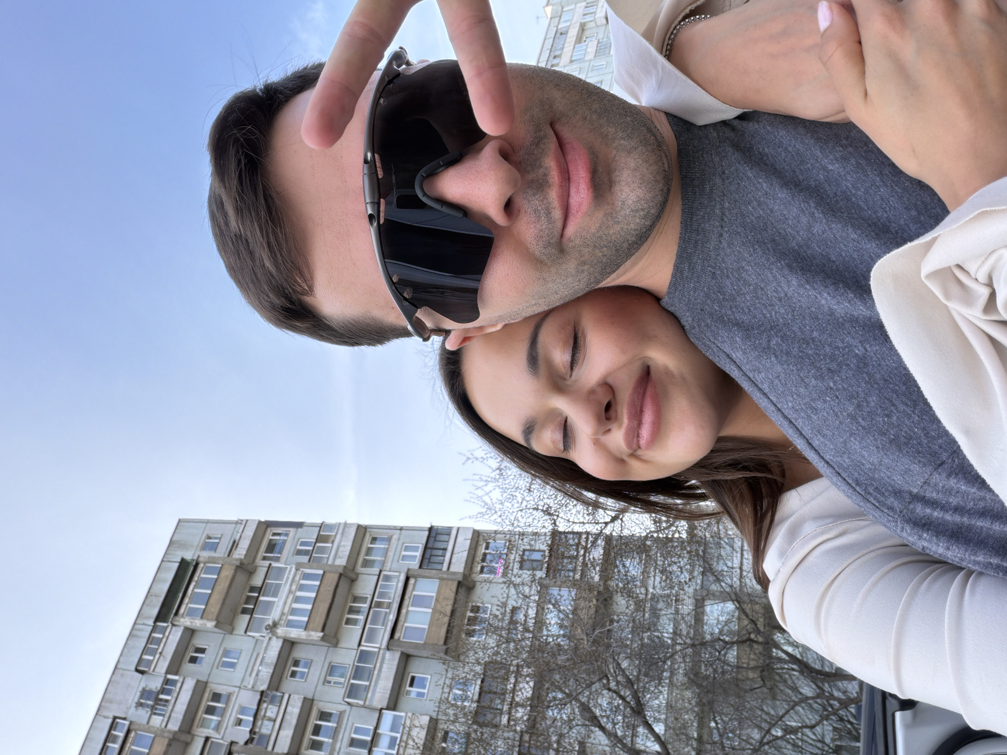 This close-up selfie captures a moment of affection between a couple in an urban environment. In the foreground, a man with dark hair and light stubble is depicted with his head slightly tilted. He wears dark, wraparound sunglasses, obscuring his eyes, and a grey t-shirt. His right hand, with two fingers raised in a V-sign, is partially visible at the top, indicating he is holding the camera.
Resting her head gently against his shoulder or chest is a woman with dark hair. Her eyes are closed, and a soft, contented smile plays on her lips, conveying a sense of peace and happiness. She is wearing a light-colored, possibly cream or white, collared shirt layered over a grey top. Her right hand, adorned with a thin bracelet and light pink fingernail polish, is visible, resting on the man's arm.
The background reveals a clear, light blue sky on the left side of the frame. On the right, a large, multi-story apartment building stands prominently, characterized by a utilitarian or Brutalist architectural style with numerous windows and concrete elements, typical of structures found in post-Soviet countries. Bare tree branches with fine twigs are visible in the midground, suggesting a season like late winter or early spring.
The lighting is soft and natural, indicating daytime. The scene evokes a feeling of intimacy and quiet joy between the subjects. The location, as provided, is Krasnoyarsk, Russia. There is no visible text in the image.