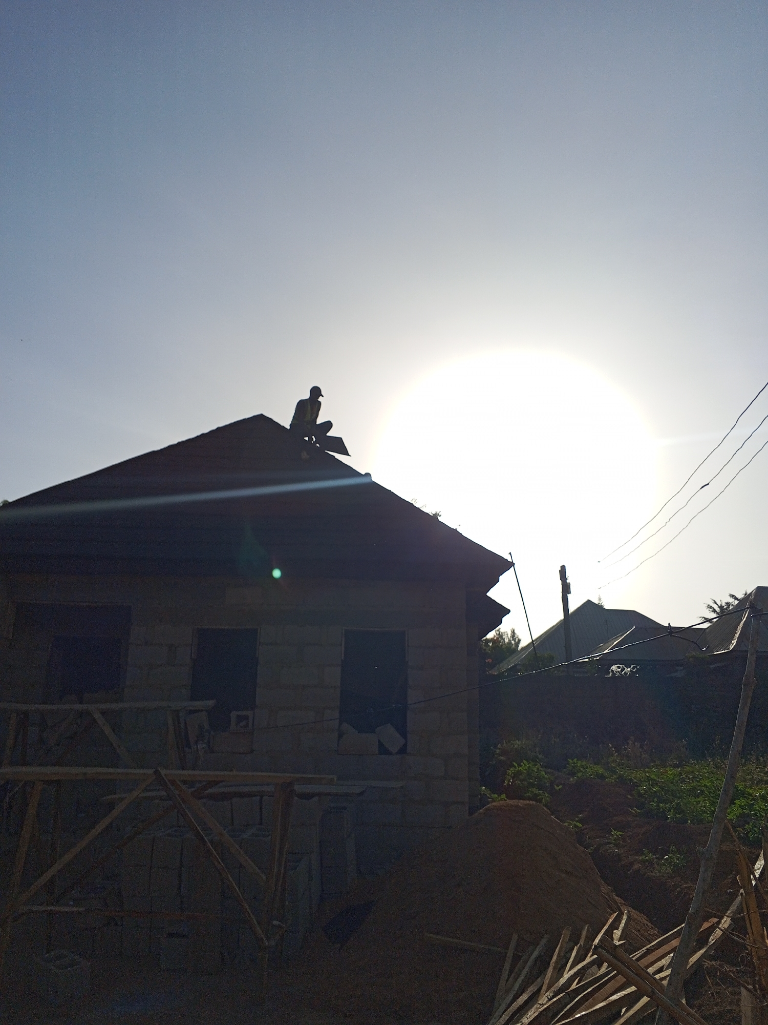 A silhouetted figure is perched on the roof of a building under construction in Bukuru, Nigeria, against a bright sky with the sun obscured by haze. The building is made of cinder blocks, with visible window openings and unfinished wooden scaffolding. In the background, another building and some vegetation are discernible. Construction materials, including a pile of sand and scattered lumber, are present in the foreground. Power lines are visible extending from the side of the house.