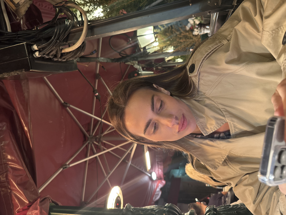A woman with long brown hair and a light complexion is seated in an outdoor dining area in Venice, Italy. She is looking downwards at a smartphone, partially visible in the foreground with a clear protective case, held in her hands. She is wearing a light tan trench coat over a plaid shirt. Above her, a dark red outdoor umbrella or canopy with visible metal ribs provides shelter. To the left, a circular infrared heater is attached to a decorative black pole. On the right, a bundle of electrical cables is secured to another dark vertical support beam. The background features multiple illuminated outdoor lanterns and potted greenery, with blurred figures of other patrons suggesting a public establishment during evening hours or in a low-light environment.