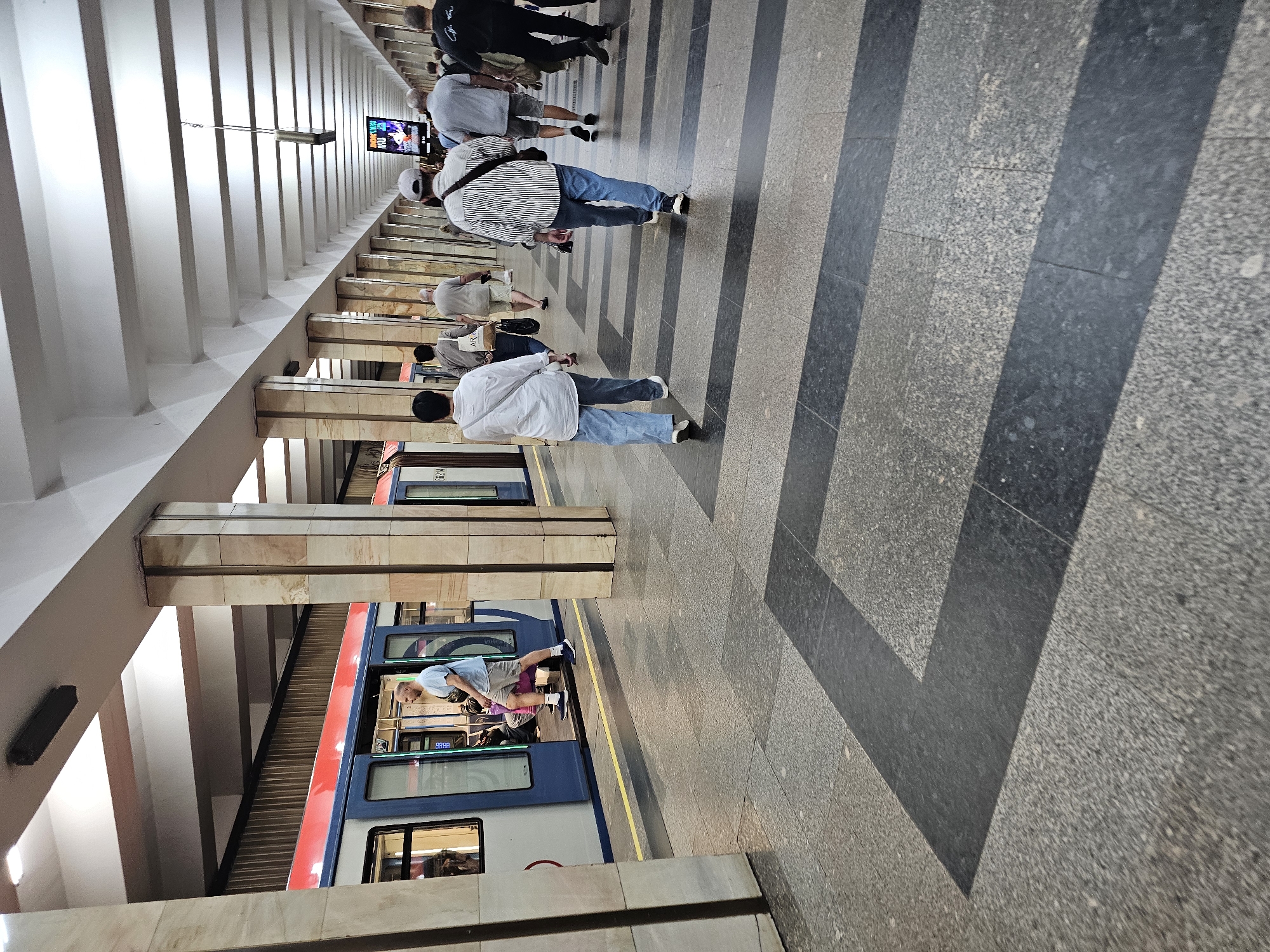 A man in a light blue t-shirt and grey shorts exits a train car onto a subway platform in Khimki, Russia. The train is red and blue. The platform is covered by a concrete ceiling supported by marble-clad pillars. The floor is tiled with granite and features a pattern of dark grey rectangles. Several other people are walking away from the train along the platform. One person in a striped shirt walks with a black bag slung over their shoulder. Another person in a white shirt and blue jeans walks in the foreground. A digital advertisement screen is visible in the distance.