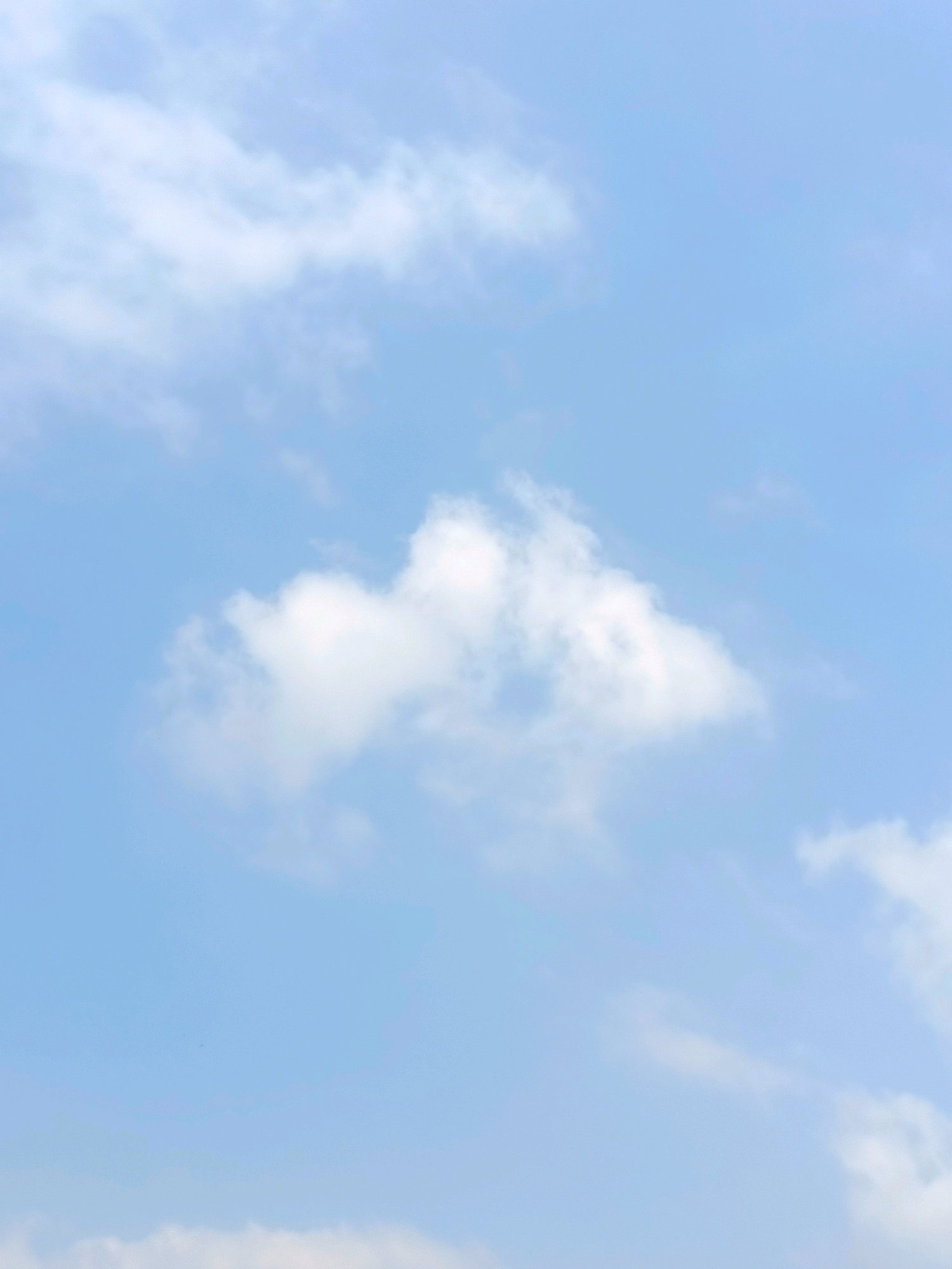 The image is a vertical shot of a light blue sky with scattered white clouds. One prominent cloud formation is centrally located in the upper half of the image. The bottom edge of the frame shows a sliver of what appears to be the edge of other clouds, suggesting the presence of a sky filled with a moderate amount of cloud cover. There are no identifiable structures, landforms, or human elements visible in this sky-only composition. The lighting suggests it is daytime.